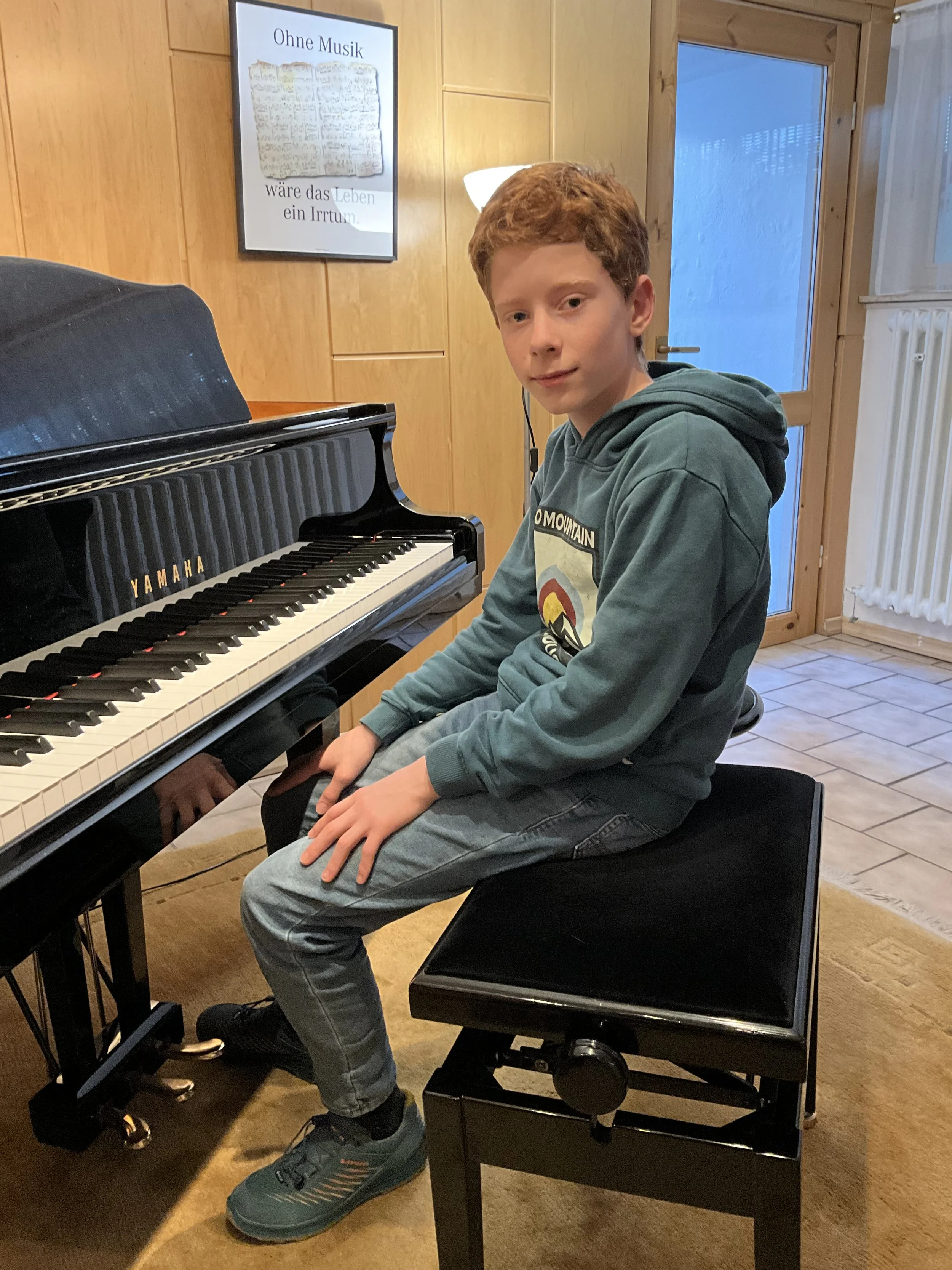 Ein junger Junge sitzt an einem schwarzen Klavier, der Raum hat Holzpaneele an den Wänden und eine Glas-Tür. An der Wand ist ein Bild mit dem Text »Ohne Musik wäre das Leben ein Irrtum.«
