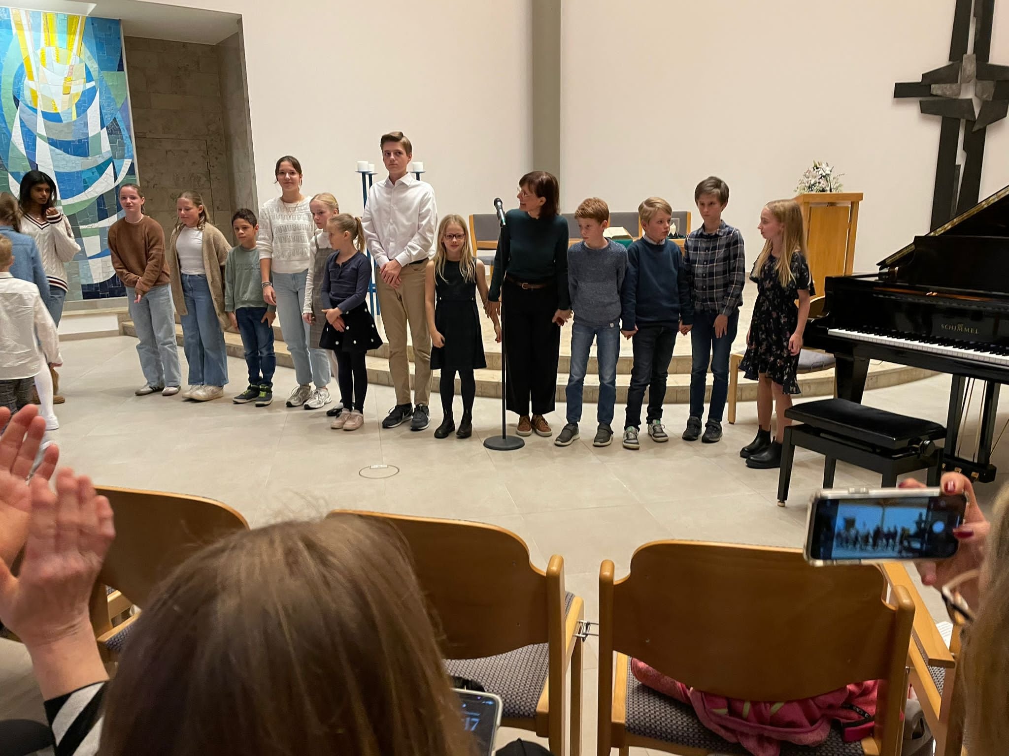 Kinder und Jugendliche stehen in einer Reihe auf einer Bühne in einer Kirche, während Erwachsene im Publikum sie fotografieren oder filmen.
