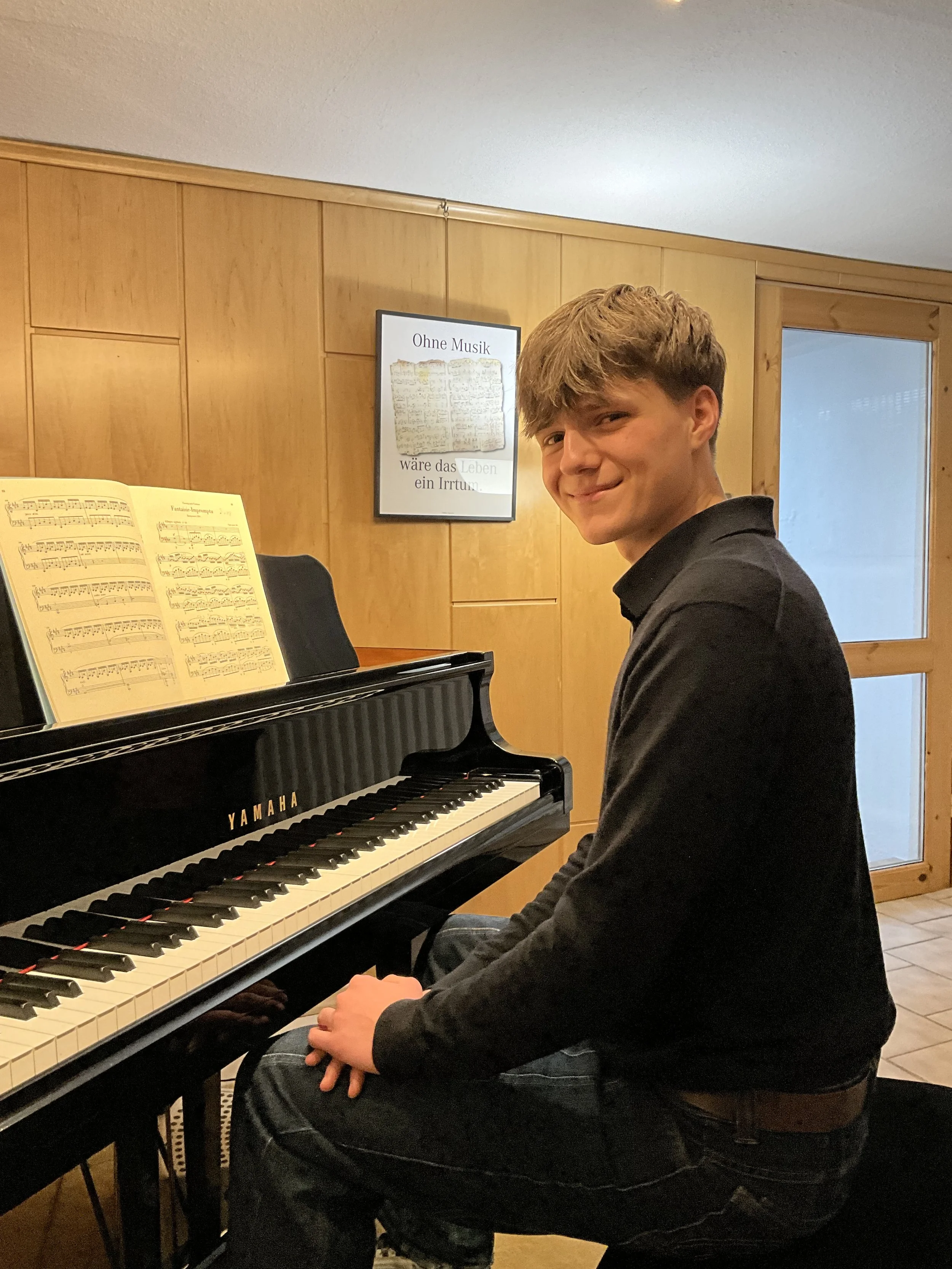 Ein junger Mann sitzt an einem schwarzen Yamaha-Flügel, spielt und lächelt. Im Hintergrund eine Holzvertäfelung und ein Bilderrahmen mit deutschem Zitat.