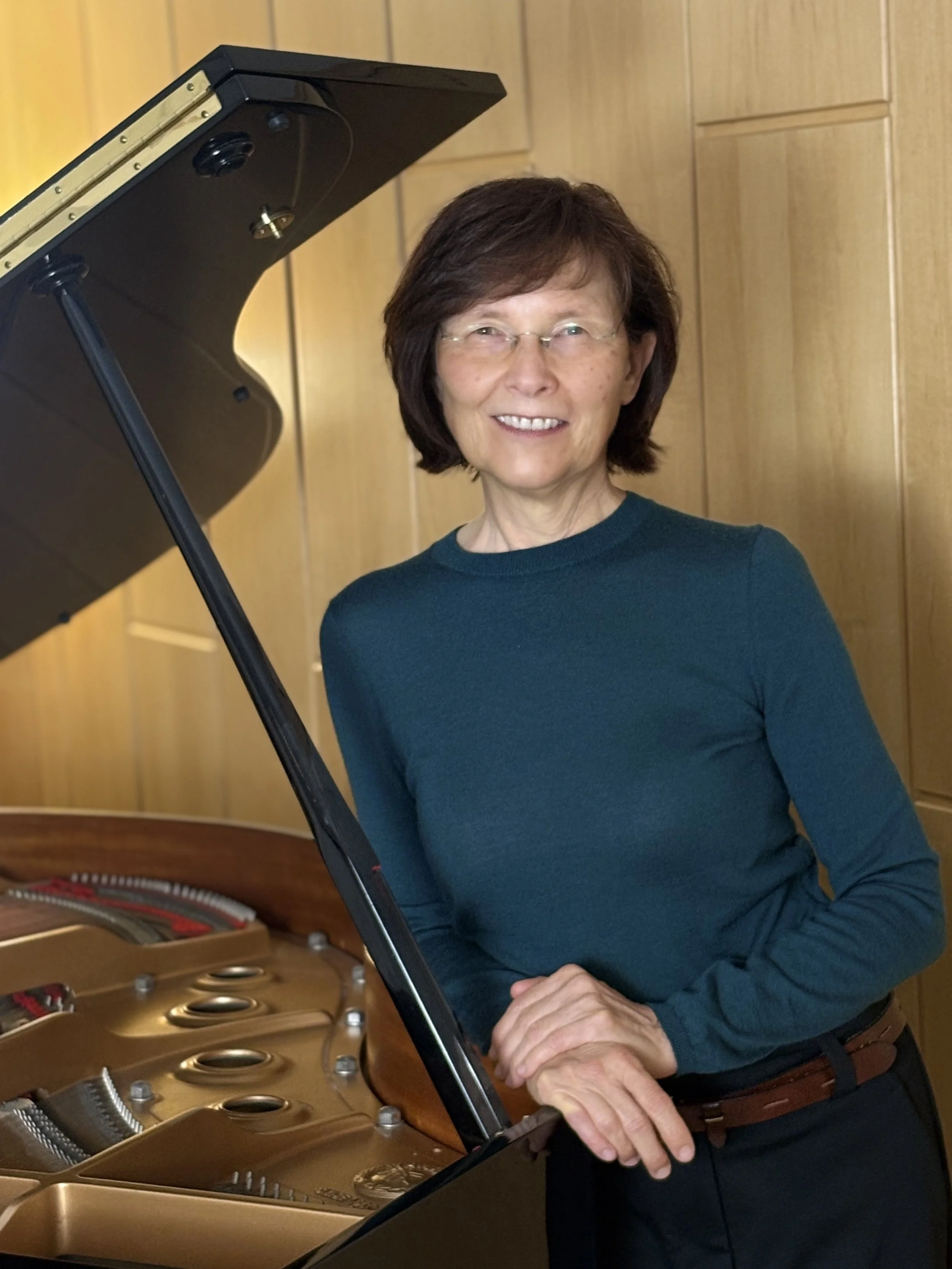 Beate May mit braunen Haaren, Brille und einem blauen Pullover steht neben einem offenen Flügel in einem Raum mit Holzwänden.
