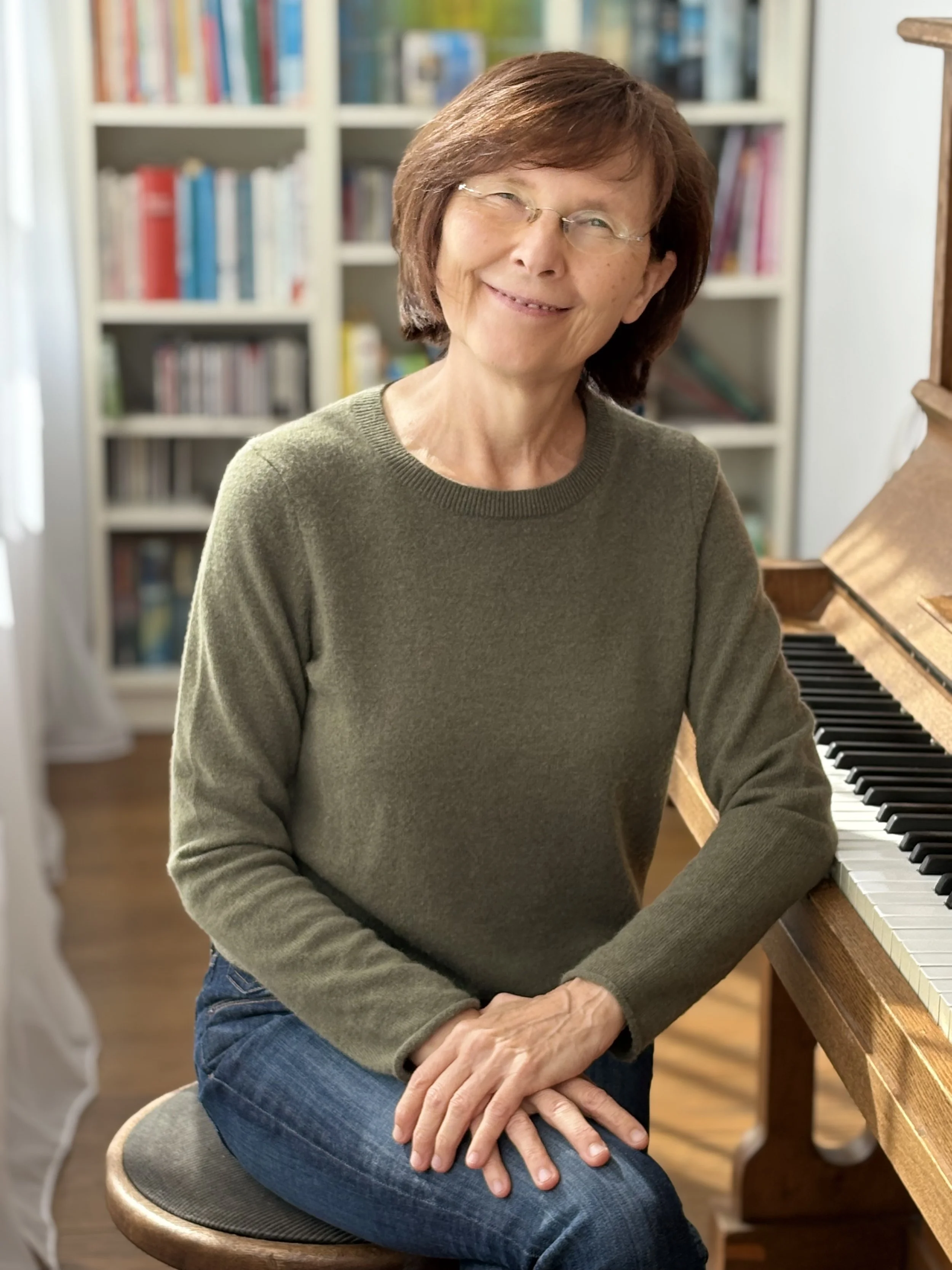 Beate May sitzt an einem Klavier, trägt eine grüne sweater und hat braune Haare. Sie lächelt in die Kamera.