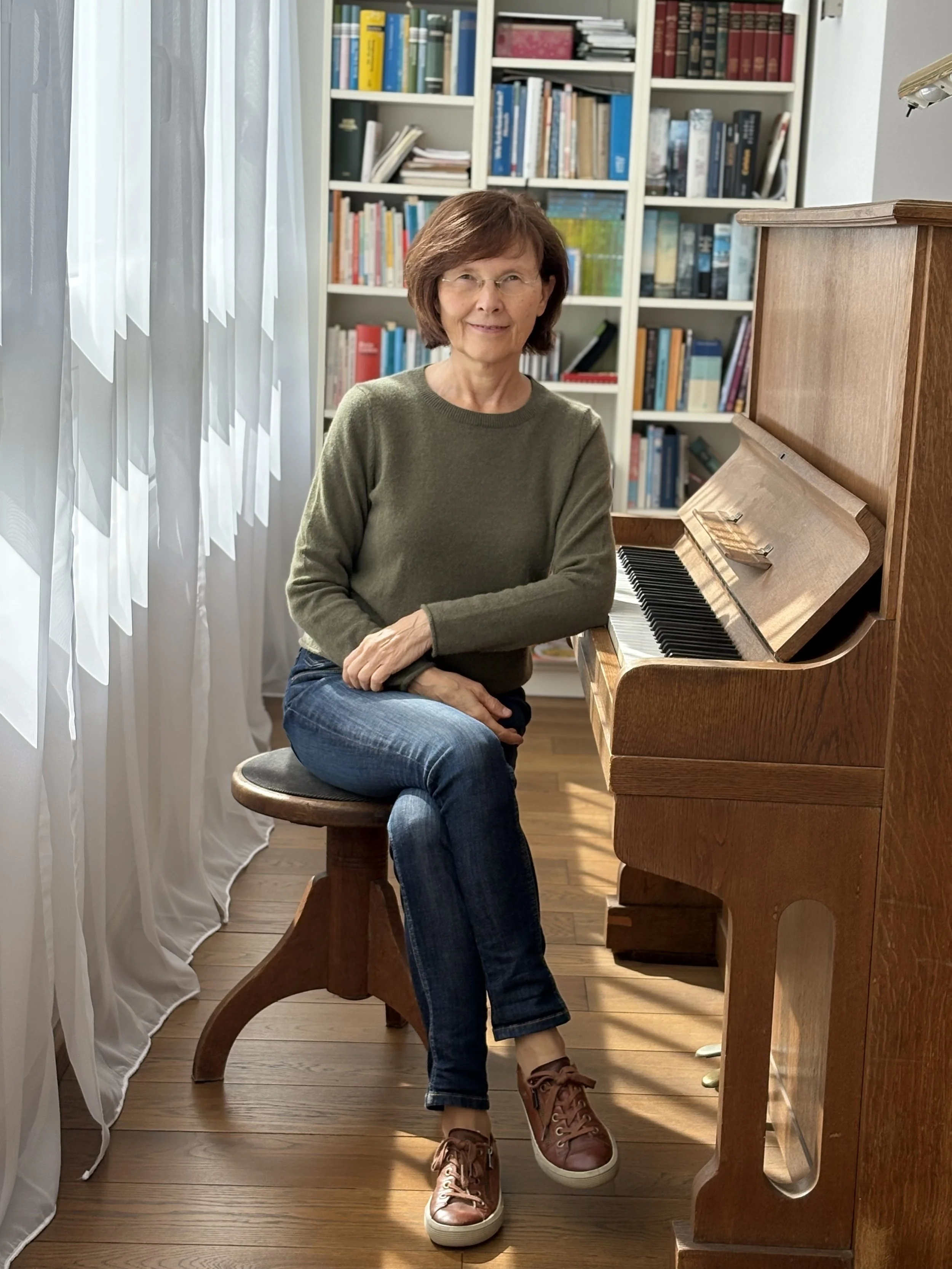 Beate May sitzt auf einem Hocker vor einem Klavier in einem Bücherzimmer.