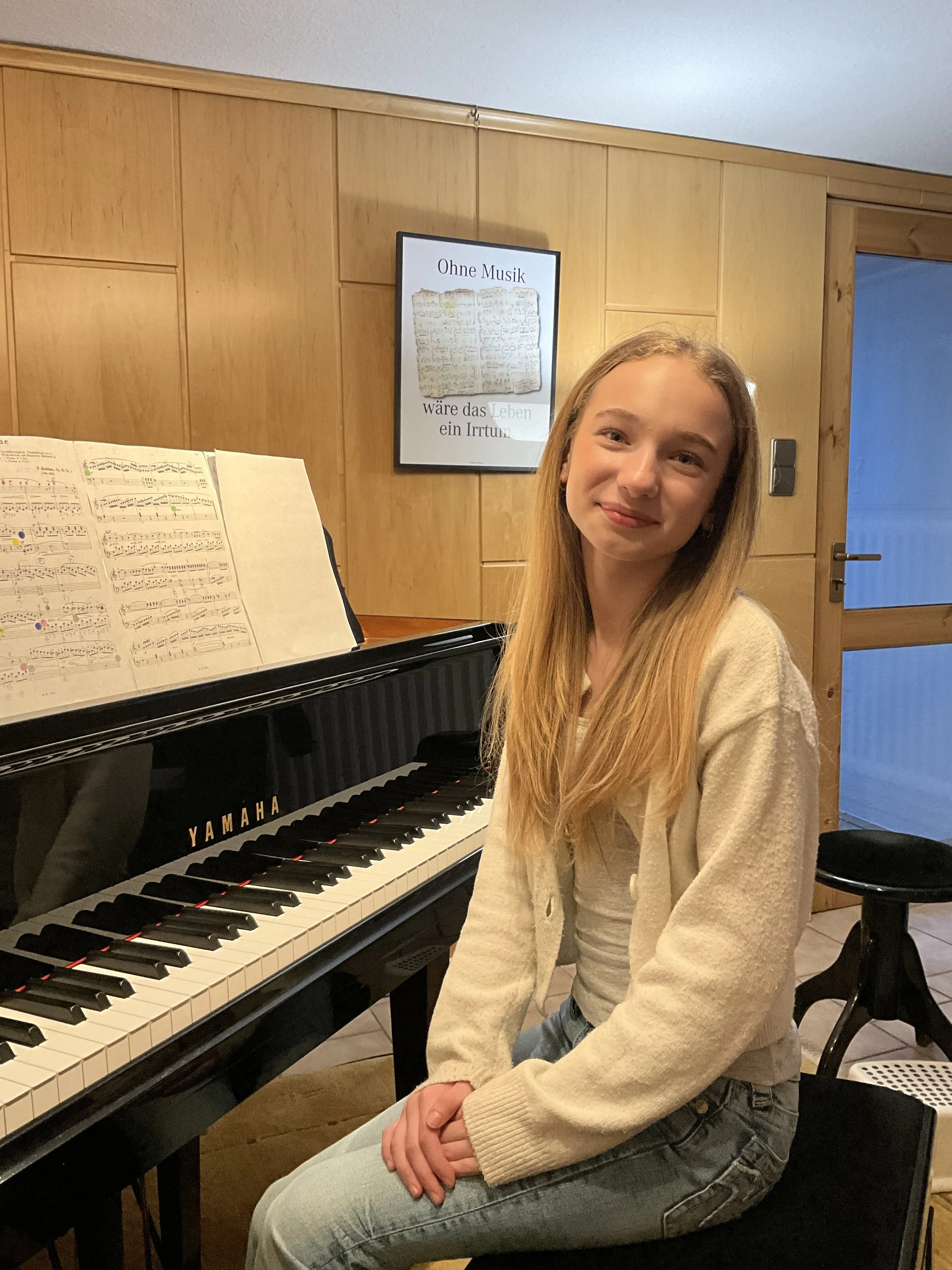Ein junges Mädchen sitzt am Klavier, lächelnd, in einem Raum mit Holzwand und einem Bild an der Wand, das Text enthält.