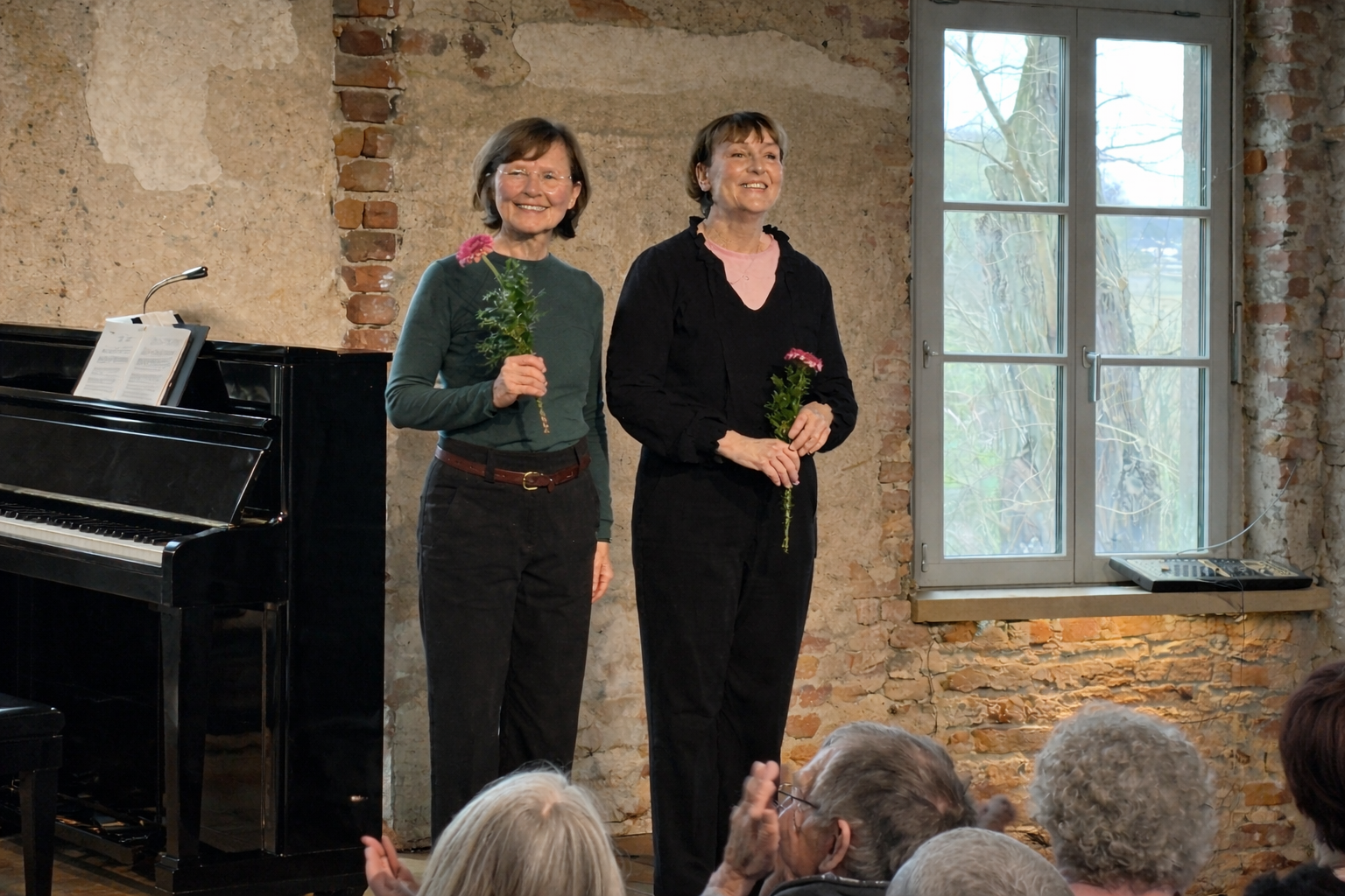 Zwei Frauen stehen vor Publikum in einem Raum mit rohen Backsteinwänden und einem Fenster, jede hält eine Blume in der Hand. Eine Frau spielt Klavier im Hintergrund.