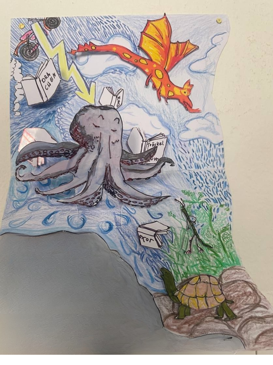 A colorful drawing featuring a flying orange dragon, an octopus with books on its head and arms, a turtle on the ground, and among green plants. The.inne is near the turtle, and there are clouds, water, and a rocky terrain depicted in the background.