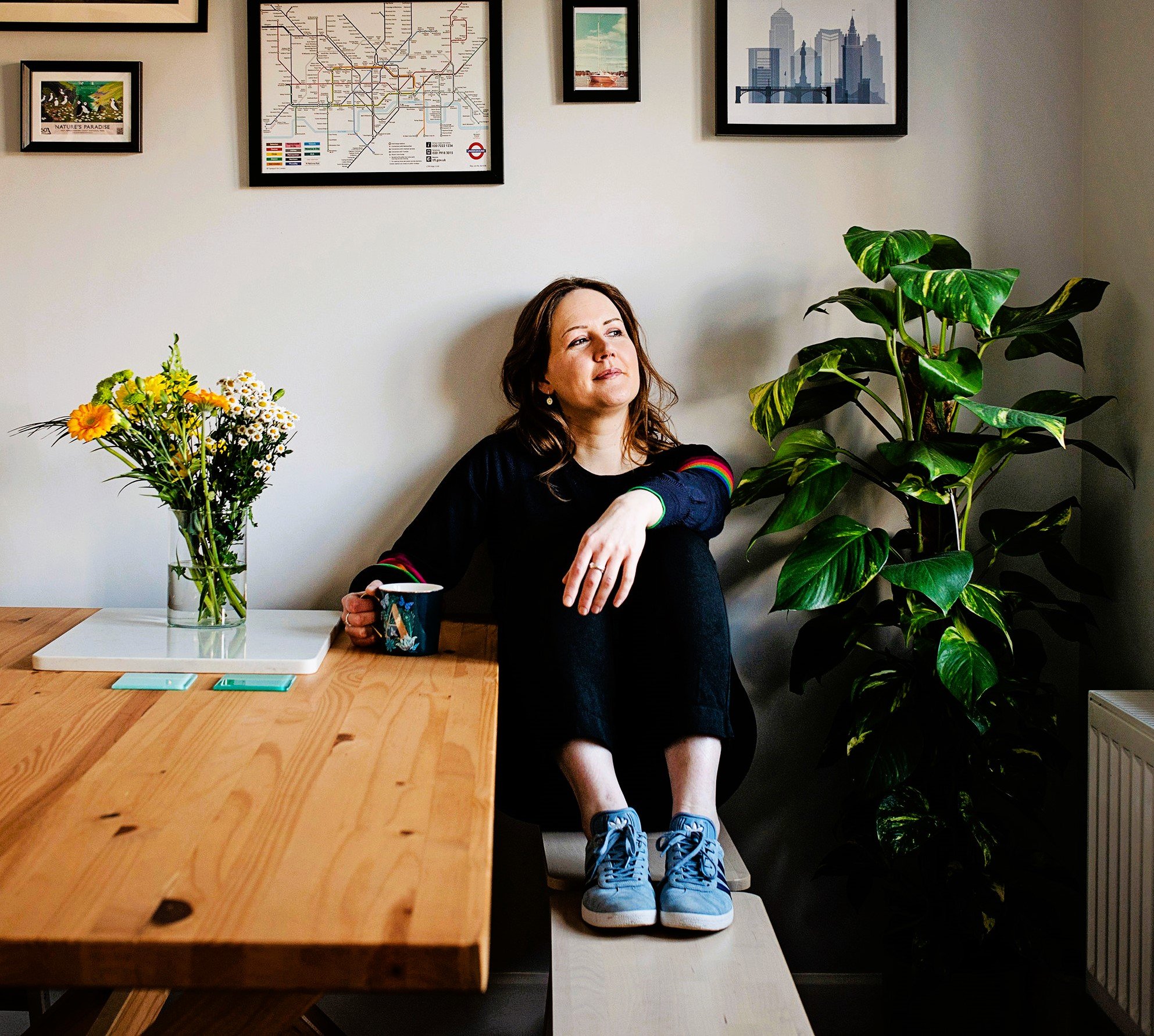 Image shows a women sitting at a table with her feet up on a bench. She is holding a mug and looking out of the window to her left. There are yellow flowers on the table and pictures on the wall behind her