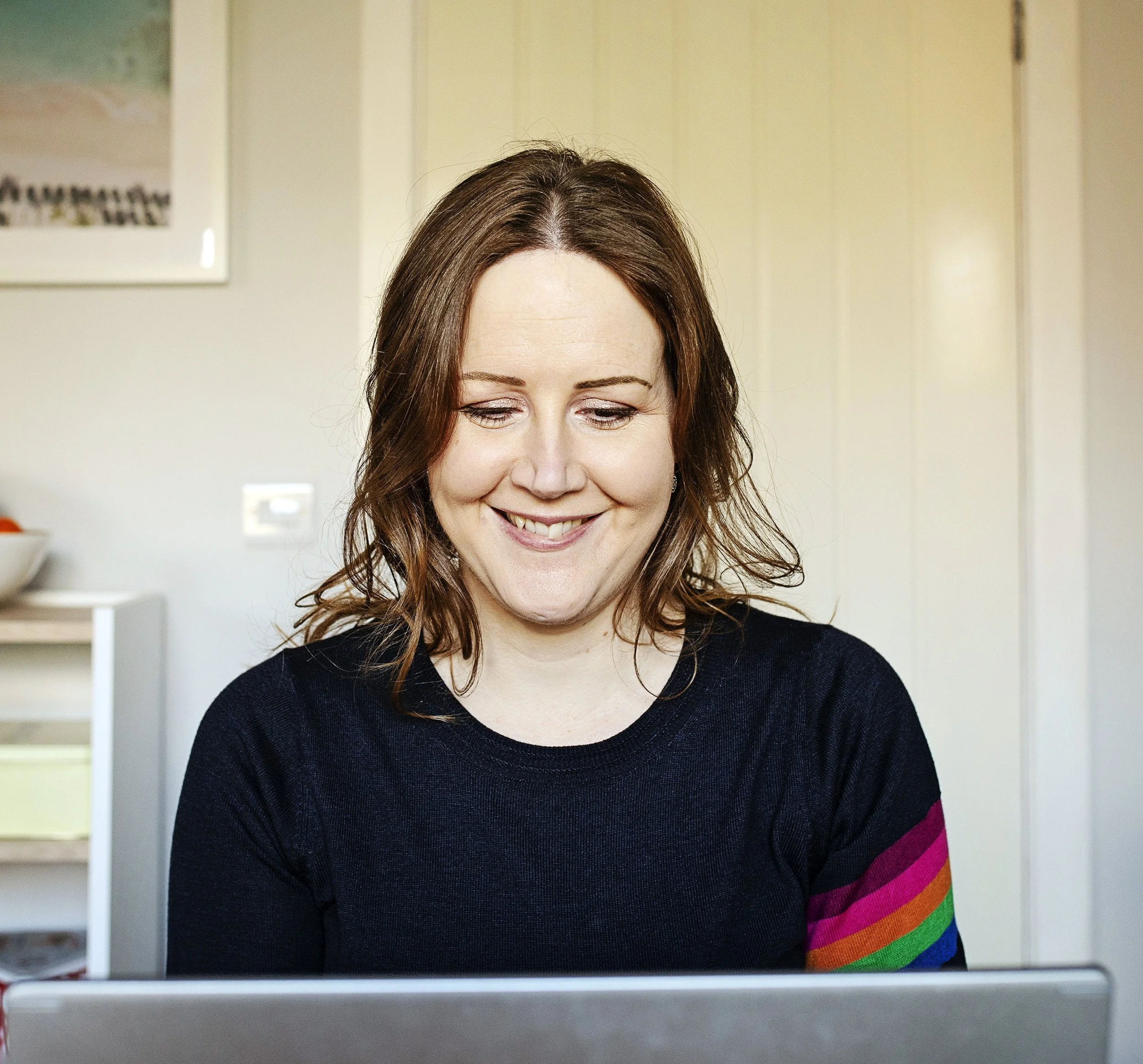 Image of a smiling woman looking down at a laptop. Behind her is a bookshelf and a closed door