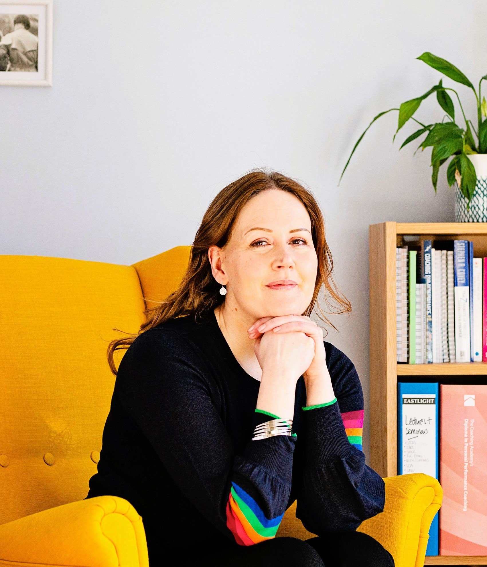 Image shows a woman in a black jumper with rainbow striped sleeves sitting in a yellow armchair. She's leaning forwards with her elbows on her knees and her chin on her hands. There is a bookcase in the background and she is smiling slightly.