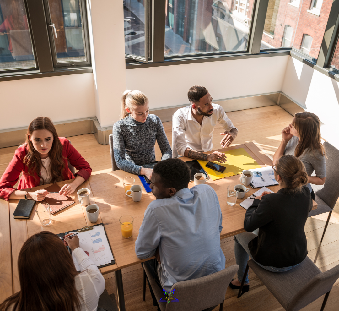 Image shows seven people sitting around an office table and talking together. On the table are water glasses, folders and files, laptops and phones. The sun is shining through the window