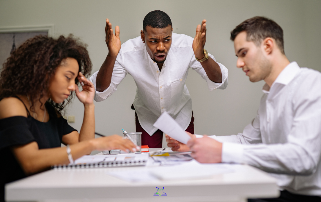 Two office workers at sat facing each other a table, looking down and avoiding eye contact. Between them on the far side of a table stands a man who is shouting at them with his hands in the air. The table is covered with papers and charts