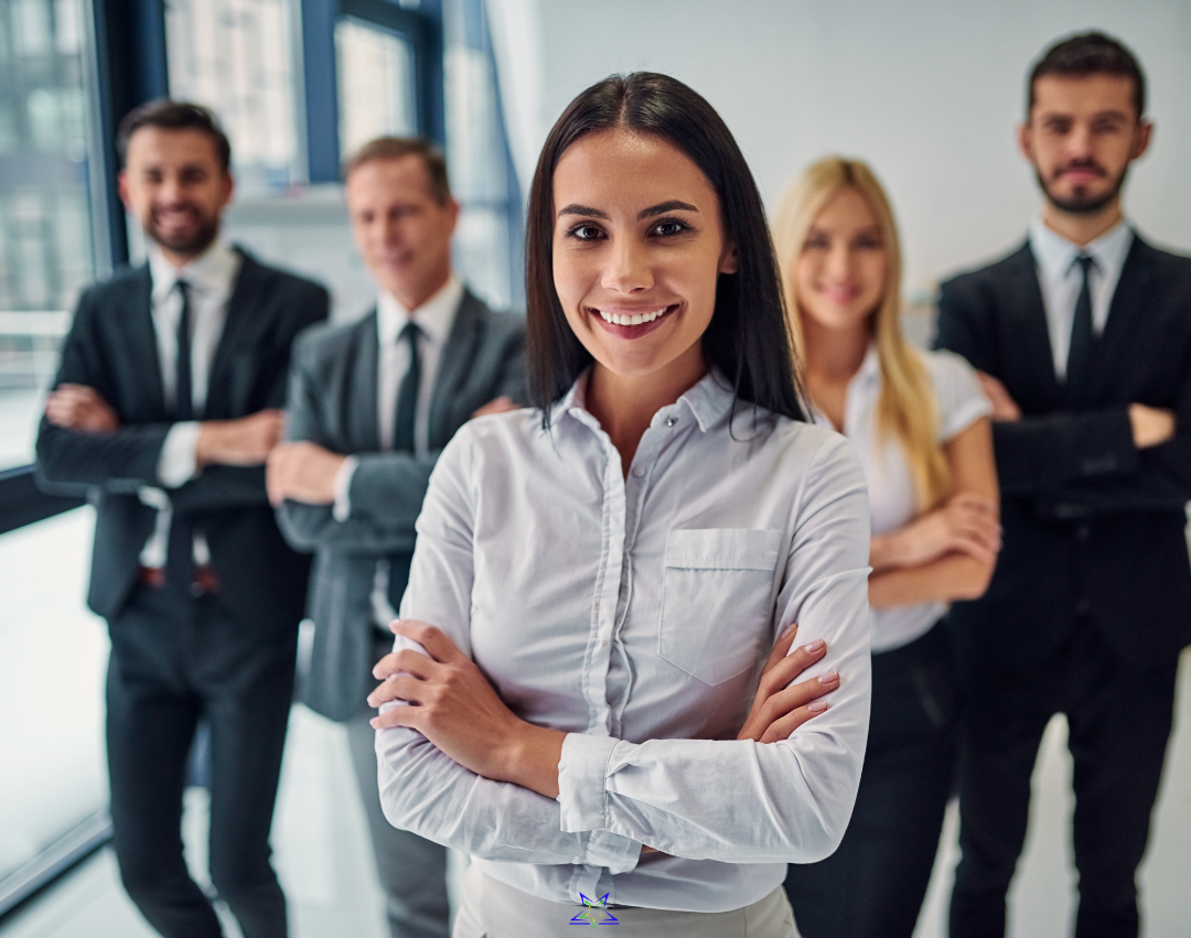 A group of five emerging leaders wearing office dress are standing smiling at the camera. There are two women and three men, all stood with their arms crossed in a bright office space