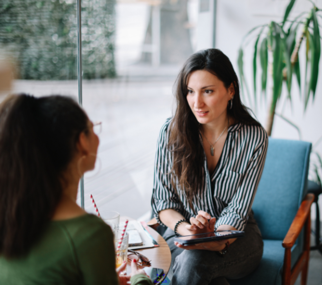 Image shows two women sitting at a table and talking. The woman on the right is wearing a striped shirt and holding a tablet. The woman on the left is wearing a green top and has her back to us. They are sitting by a large window