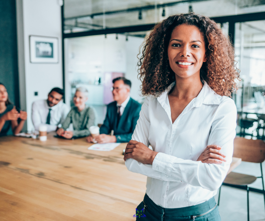 Image shows a curly haired woman in a white shirt standing in the foreground. Behind her is a large meeting table with four people sat at the table with coffee cups and paper in front of them