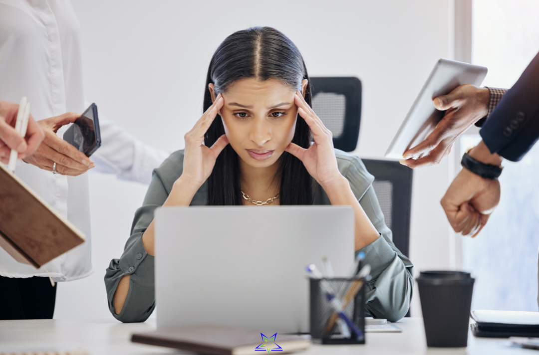 Image shows an overwhelmed woman sitting at on office desk with her fingers on her temples as the people around her try and give her a phone, notebook, reports and files