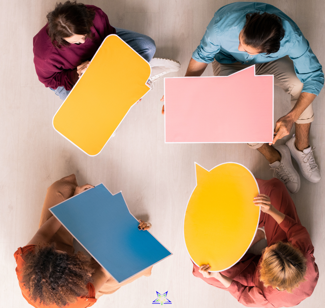 We are looking down at four people sitting in a circle on the floor. They are facing each other and each holding a differently coloured and shaped speech bubble. They are wearing casual dress and sitting on a light wood floor