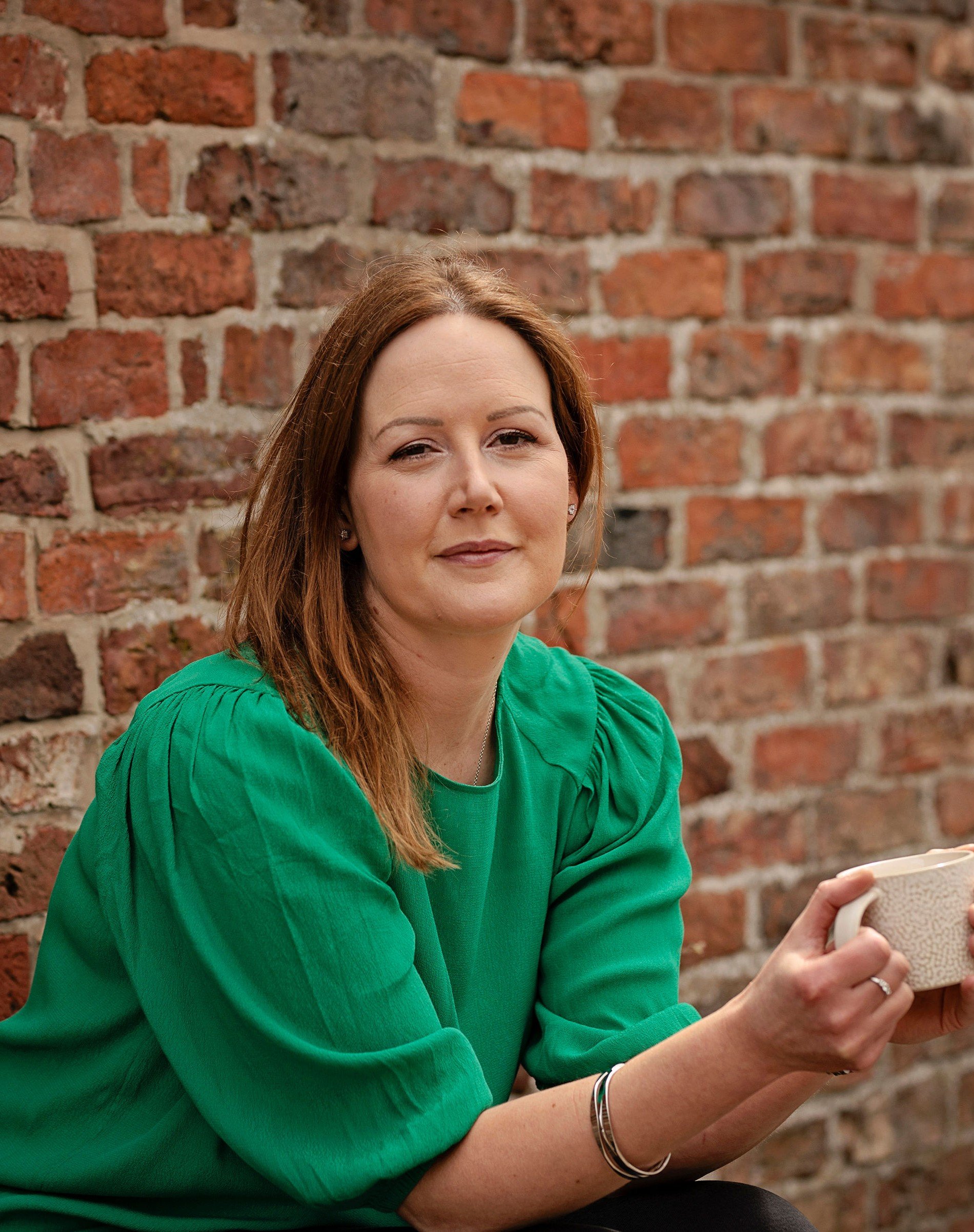 A woman wearing a green top and holding a cream mug is sitting in front of a red brick wall. She is looking at the camera, smiling slightly and squinting at the sun