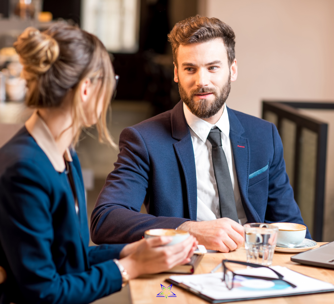 Image shows a man and a woman in office dress, sitting at a table and talking. On the table are coffee cups, files and notepads and a pair of glasses