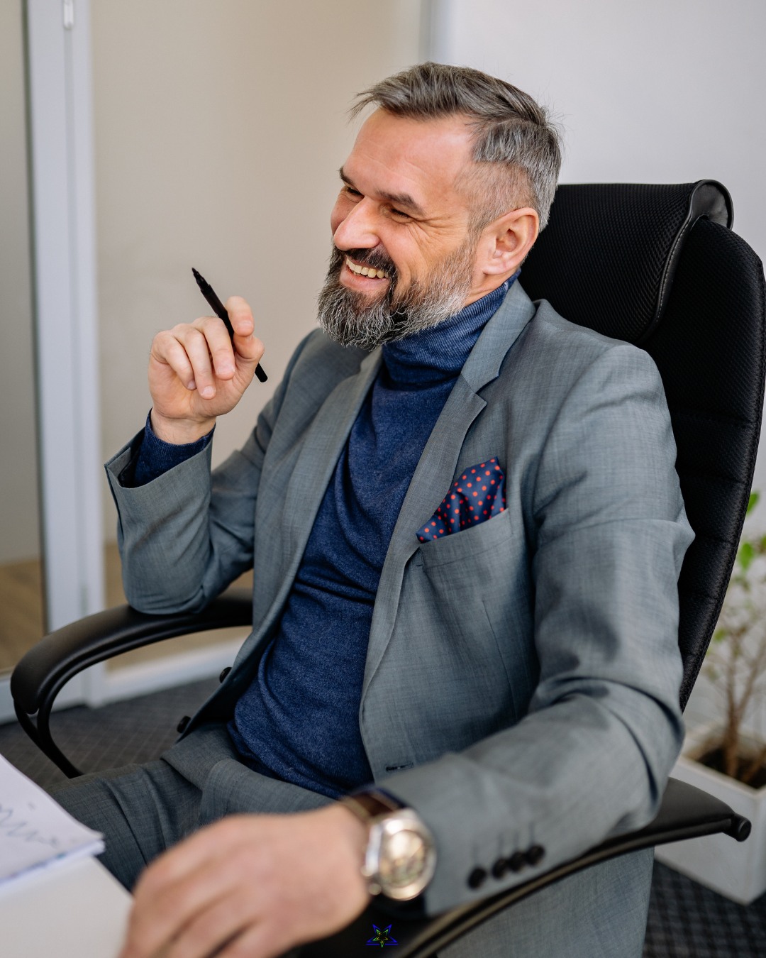Image shows a man in a grey business suit sitting at a desk. He is smiling and holding a pen in his hand
