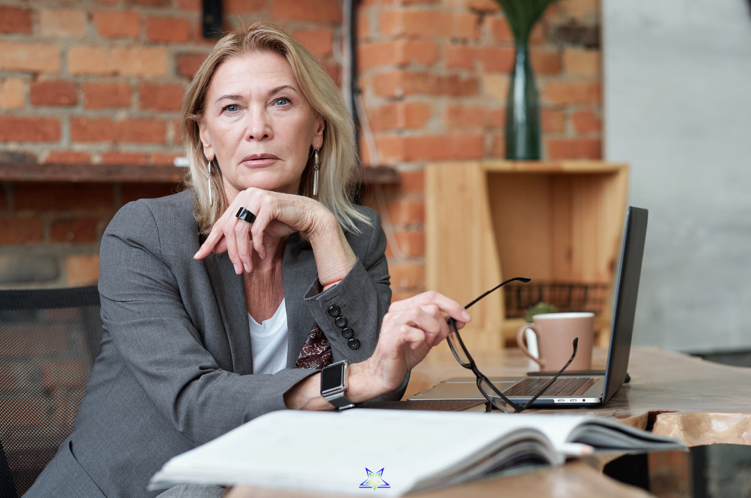 A blond haired woman in a grey business suit sits at her desk, half turned to face us. She is holding her glasses in one hand and resting her chin on her other hand. On her desk is her laptop, notebook and coffee cup. Shelves are on the brick wall
