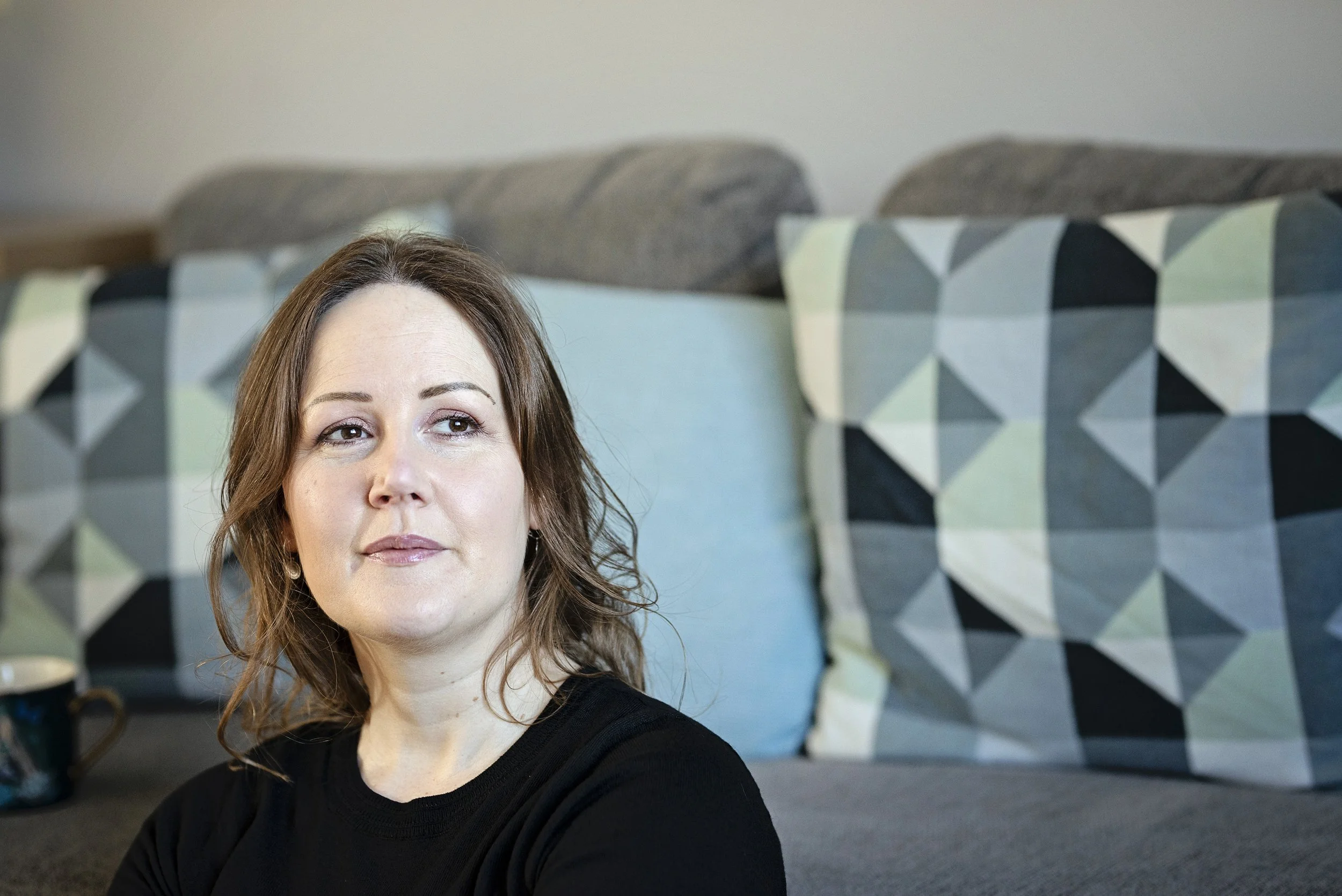 Image shows a woman in a black jumper sitting in front of a grey sofa. There are teal and blue cushions on the sofa and the woman is looking up to the right