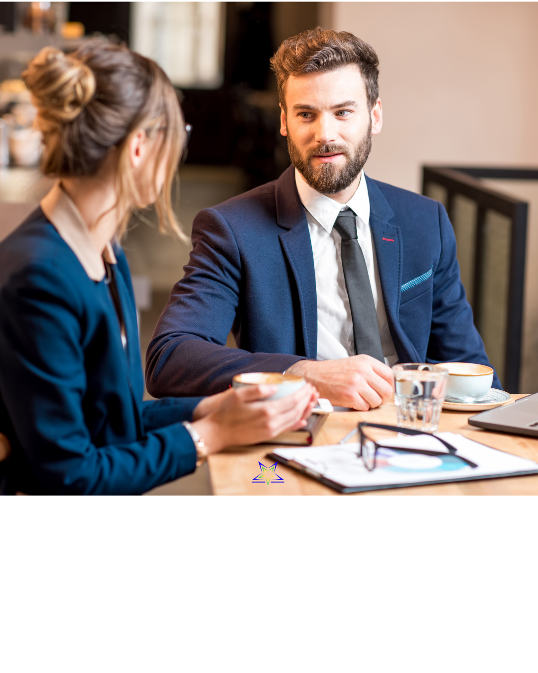 Image shows a man and a women in office dress sitting at a table having coffee. On the table in front of them are files, a laptop and a pair of glasses