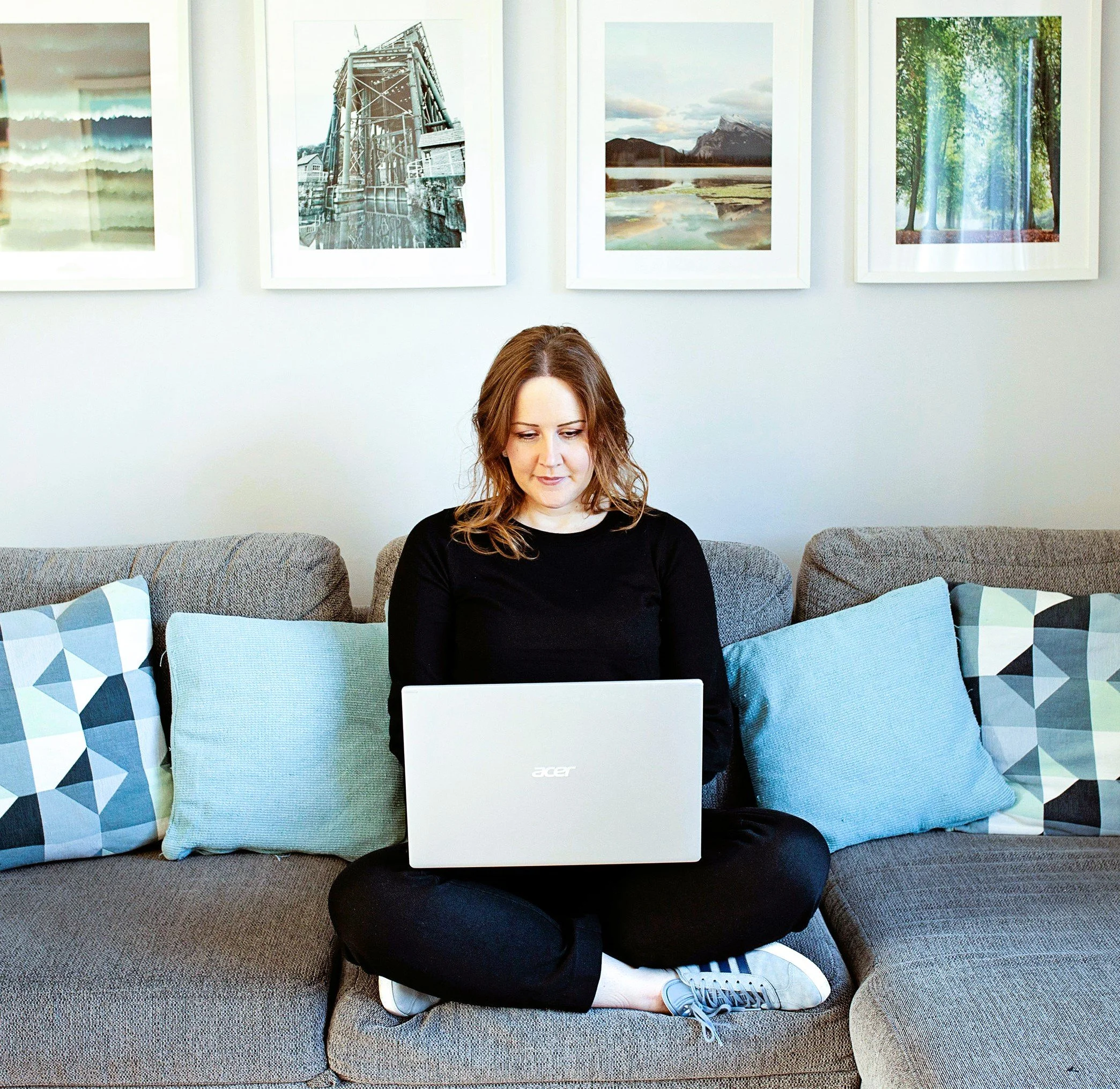 Image shows a woman in black jumper and trousers and blue trainers sitting cross legged on a grey sofa and looking down at a laptop on her lap. There are blue cushions on the sofa and four landscape pictures on the wall behind her