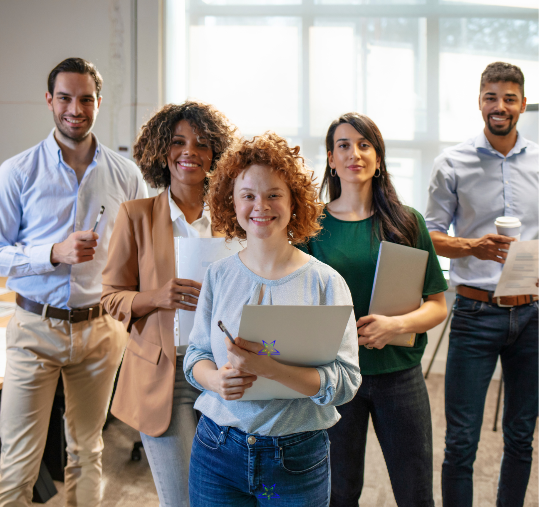 Five office workers are standing in a group, looking at the camera. The are relaxed, smiling and holding pens, files, papers and coffee cups, There are three women in the foreground and two men at the back