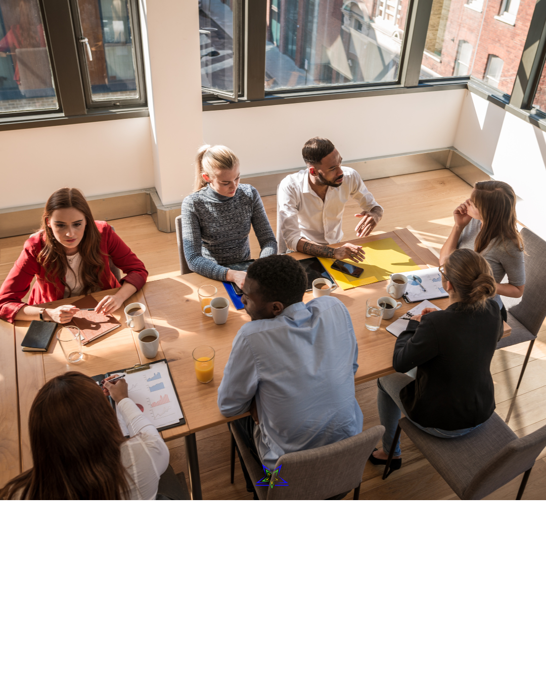 Image shows seven co-workers sitting around an office table, talking and working together. On the table there are drinks, notebooks, folders and charts. The sun shines in through the windows