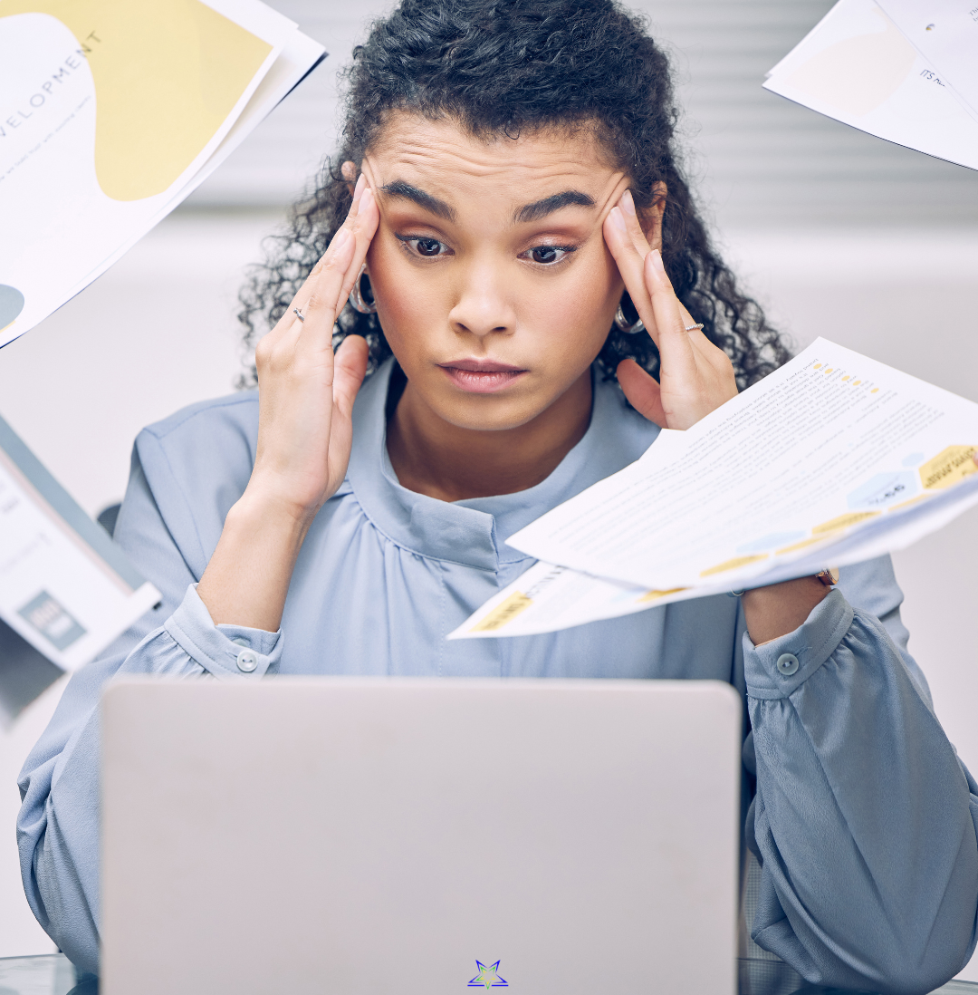 Image shows a woman rubbing her temples and looking overwhelmed as she is sat in front of her laptop and multiple people hand her reports and pieces of paper