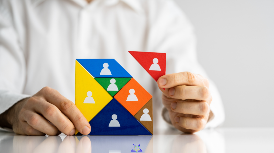 Image shows a man in a white shirt building a square puzzle. The puzzle pieces are differently coloured and sized geometric pieces, each with a white stylised head and shoulders silhoutte on