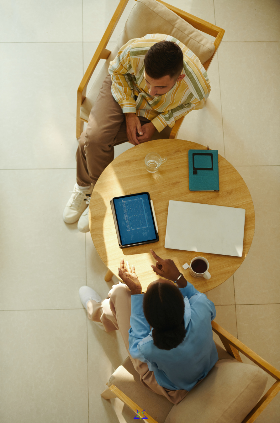 Photo is taken from above, looking down at a man and a woman sitting at a round coffee table. They are leaning forward in conversation. On the table is a tablet, laptop, notebook, water glass and tea cup. The image is light and warm