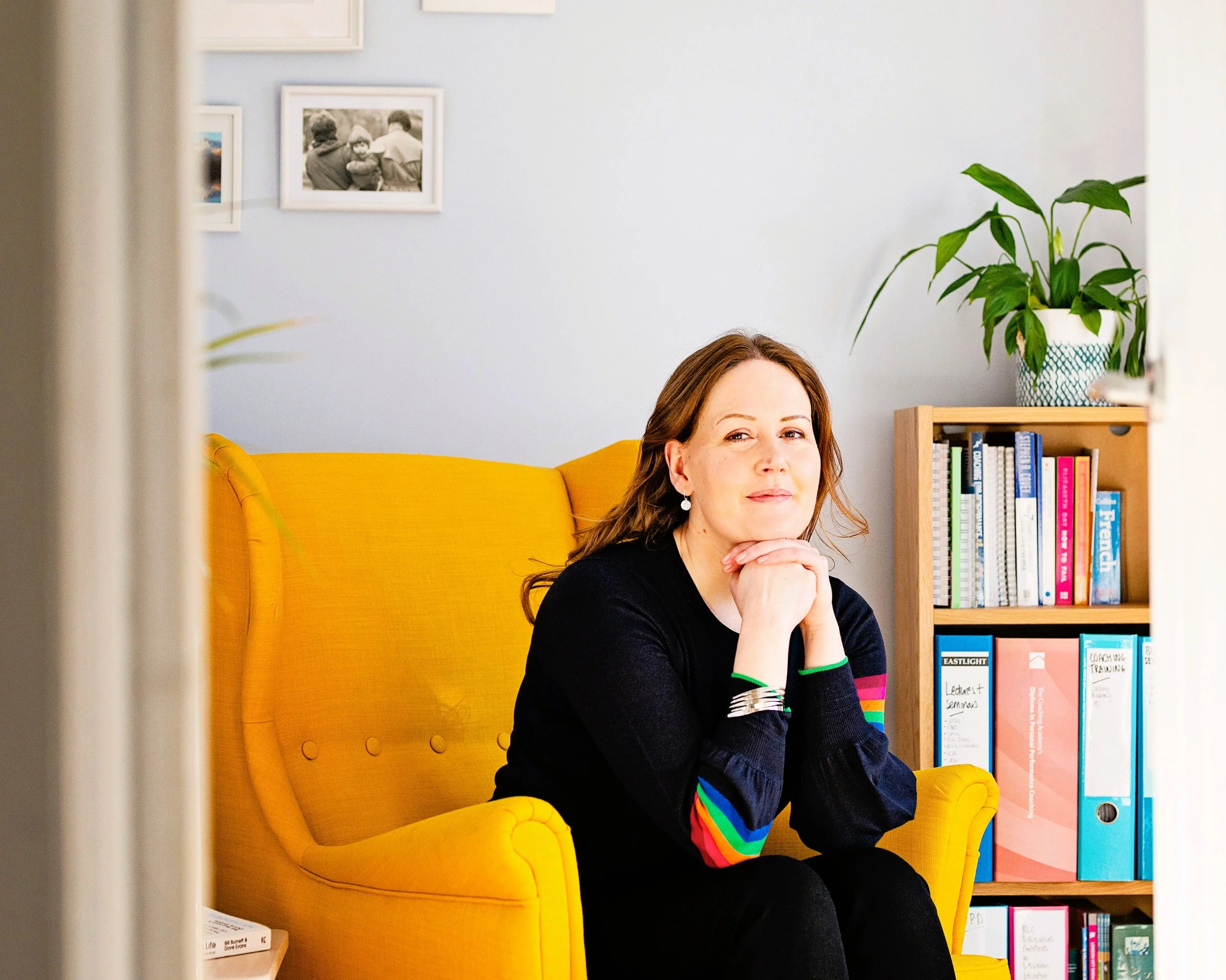 Image shows a woman in a black jumper with rainbow striped sleeves sitting in a yellow armchair with her chin on her hands. There is a bookcase full of books and files to her left and pictures on the wall behind her