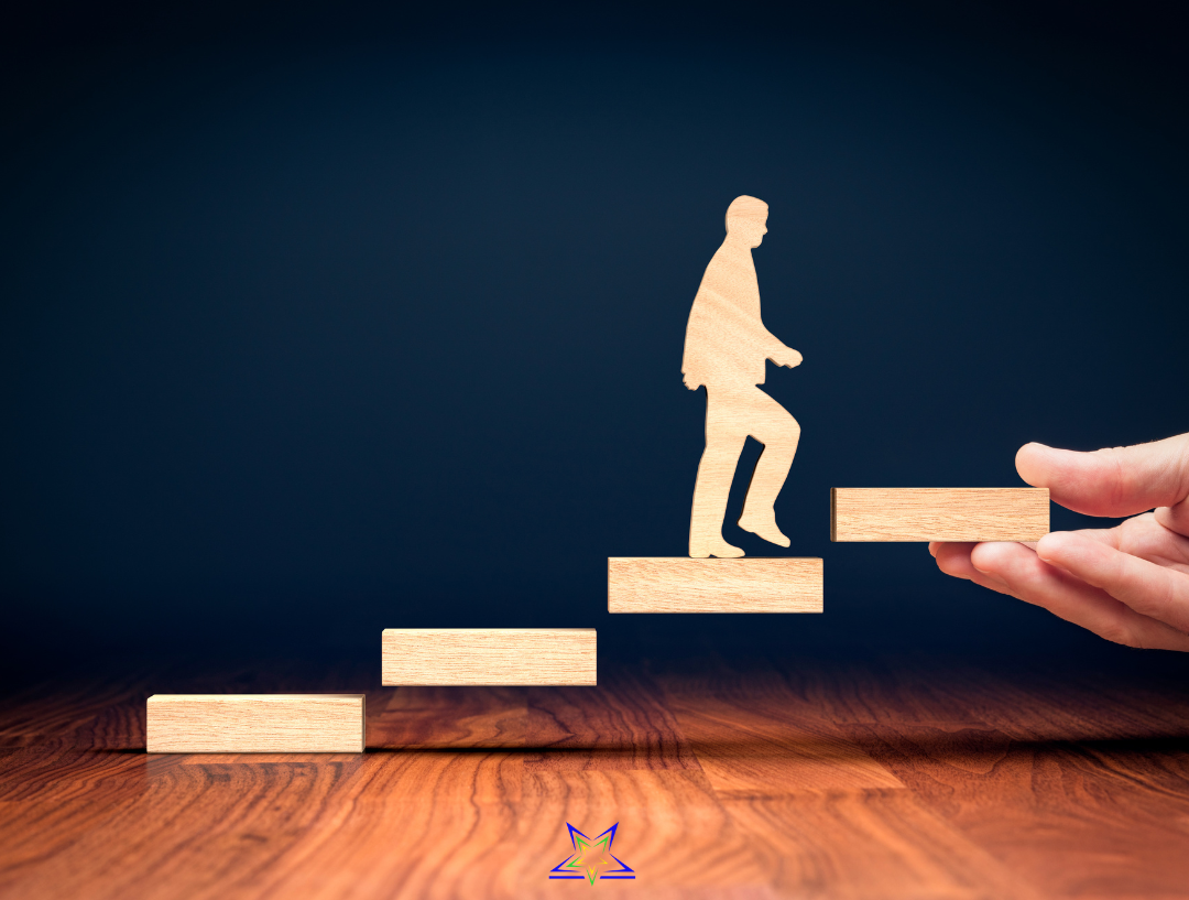Image shows four floating wooden blocks forming an ascending staircase on a wooden floor against a dark blue background. The top block is being held in a person's hand and a wooden silhouette figure is about to step up onto it