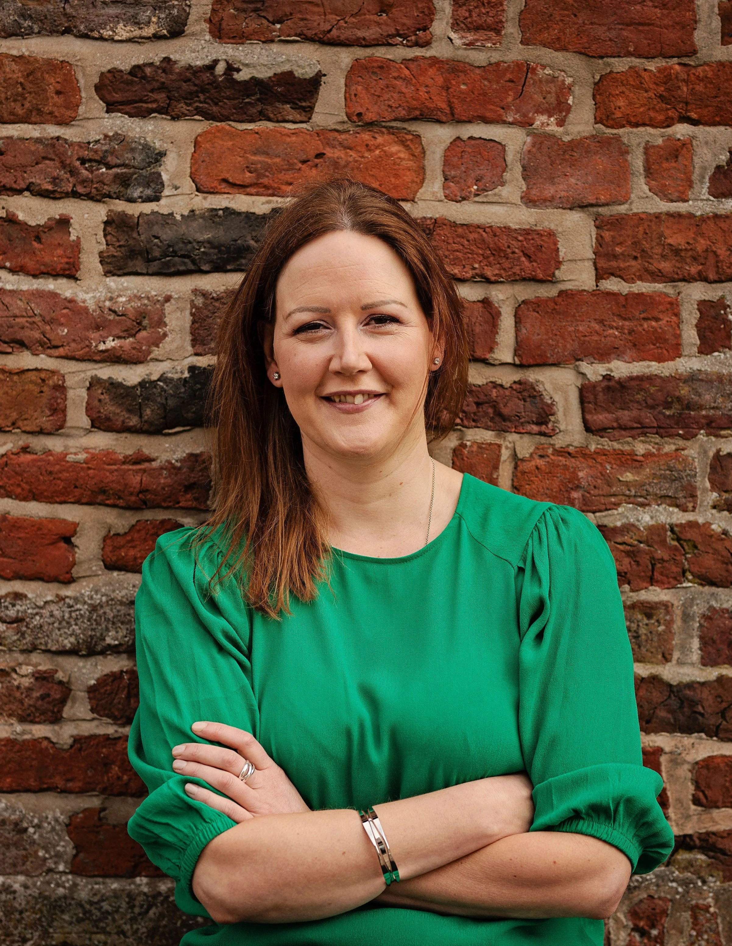 Image shows a woman in a green top and silver bracelets standing with her arms crossed against a red brick wall. She is smiling and squinting slightly against the sun