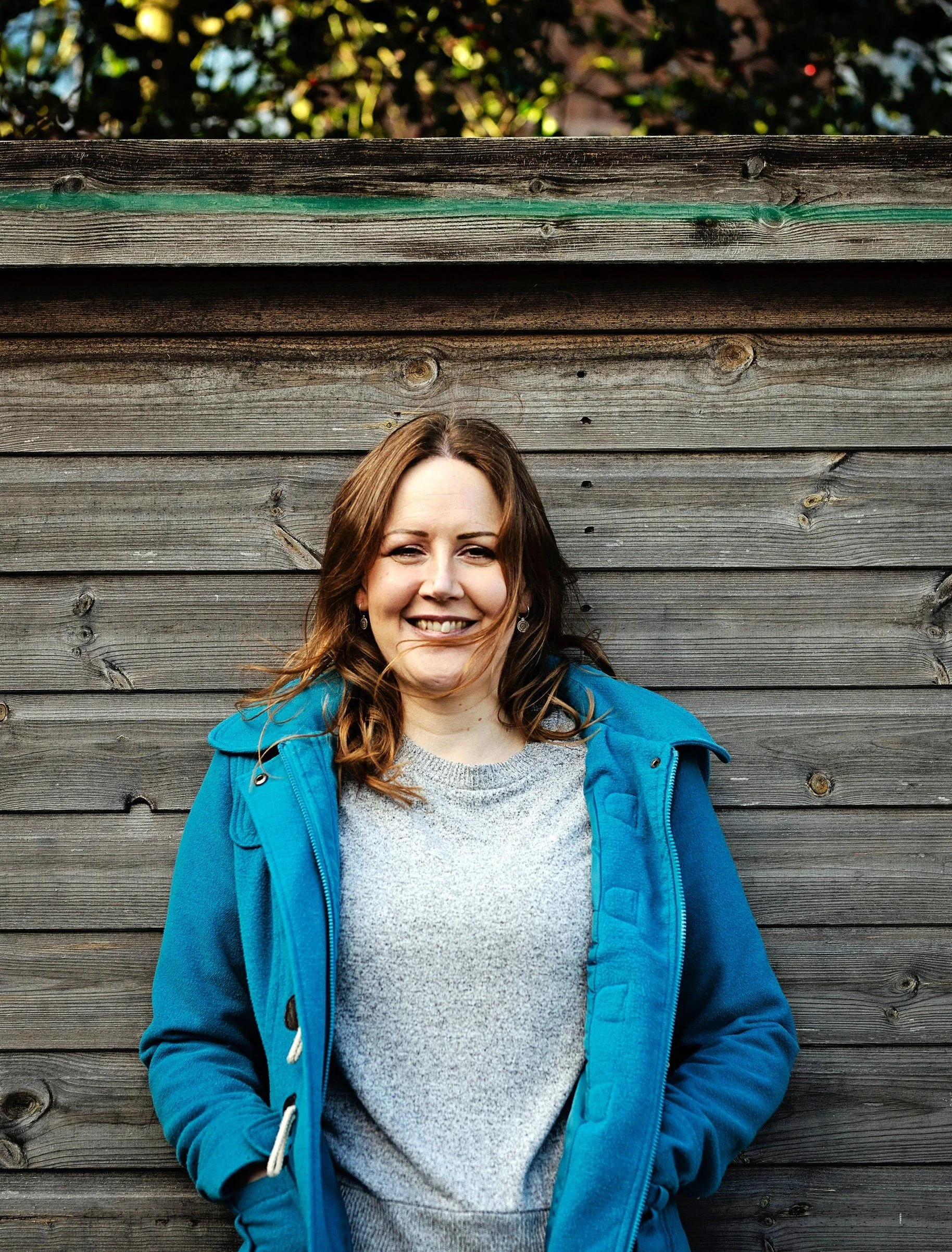 Image shows a woman in a grey jumper and blue coat standing against a wooden fence in the sunshine. Her hair is blowing across her face