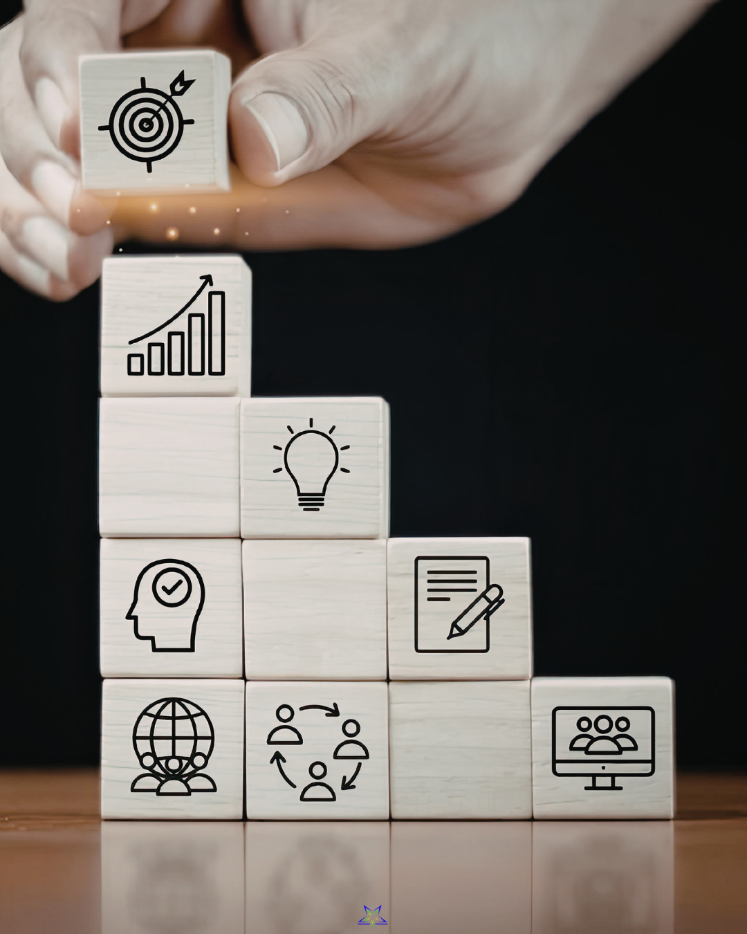 Image shows a hand building stack of square wooden blocks. The stack is four blocks wide at the bottom,decreasing by one block each layer. Some of the blocks have line drawings on them of lightbulbs, bar charts, globes, teams and heads