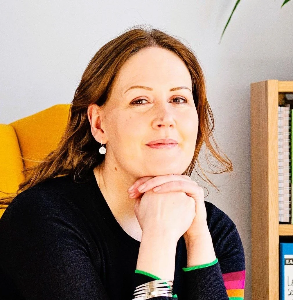 Image shows a woman in a black jumper with colourful striped sleeves sitting in a yellow armchair. She is resting her chin on her hands and smiling slightly. The edge of a bookcase can be seen in the background