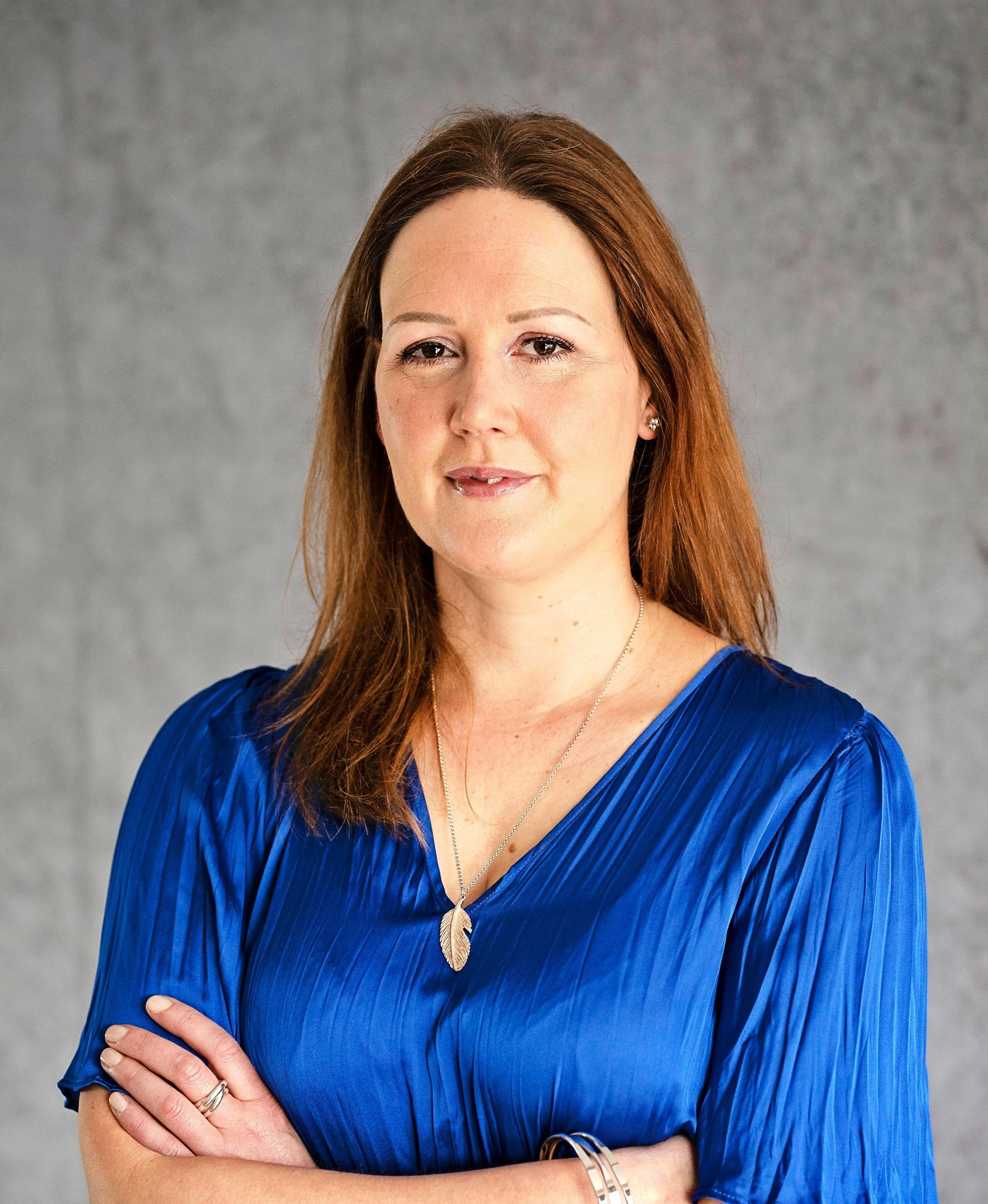 Image shows a woman in a blue top standing against a grey background with her arms crossed. She is wearing a silver leaf necklace and silver bracelets