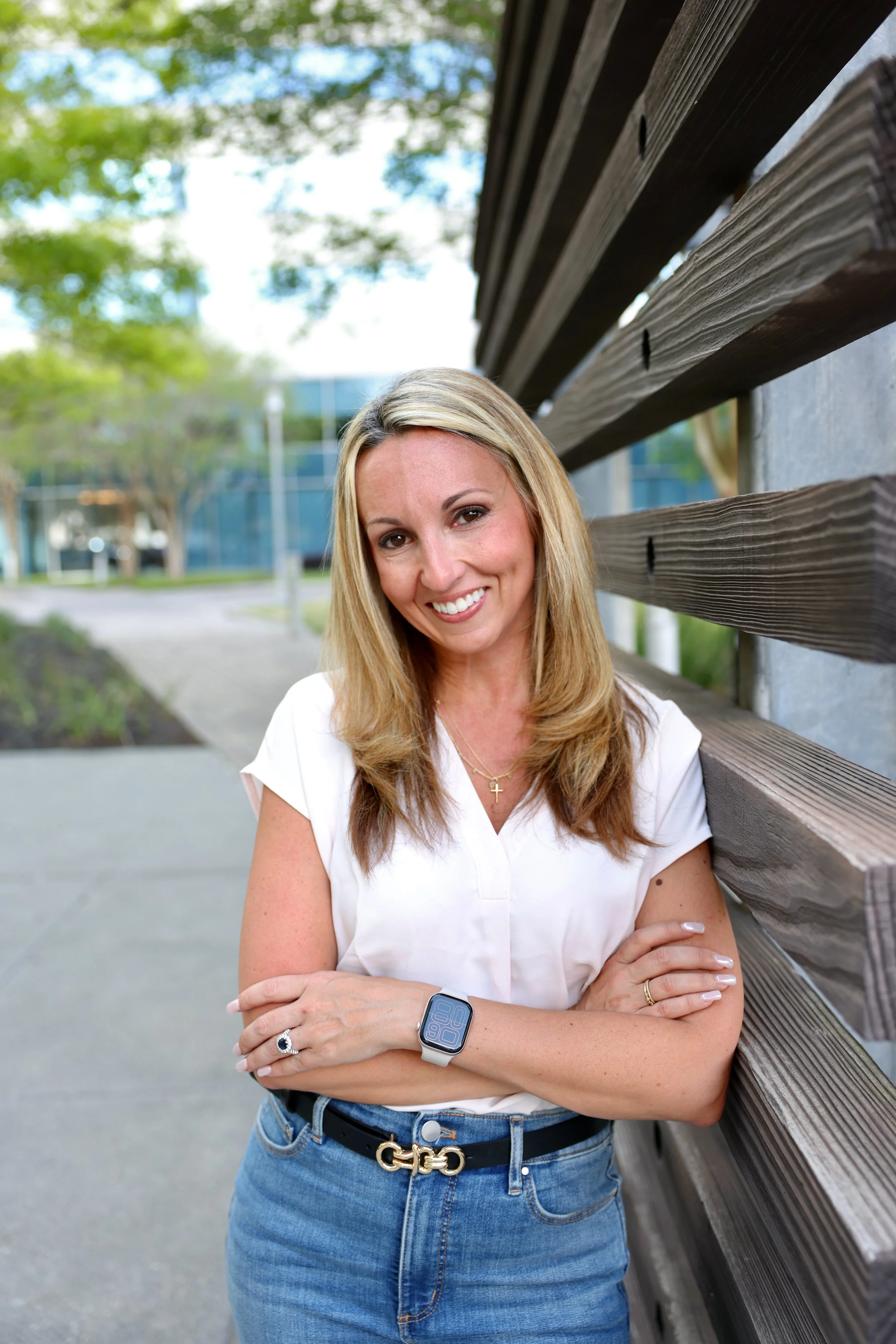 A smiling woman leaning against a wooden bench outdoors on a sunny day. She has blonde hair, wearing a white shirt, blue jeans, a gold belt, a ring, a necklace, and a smartwatch.