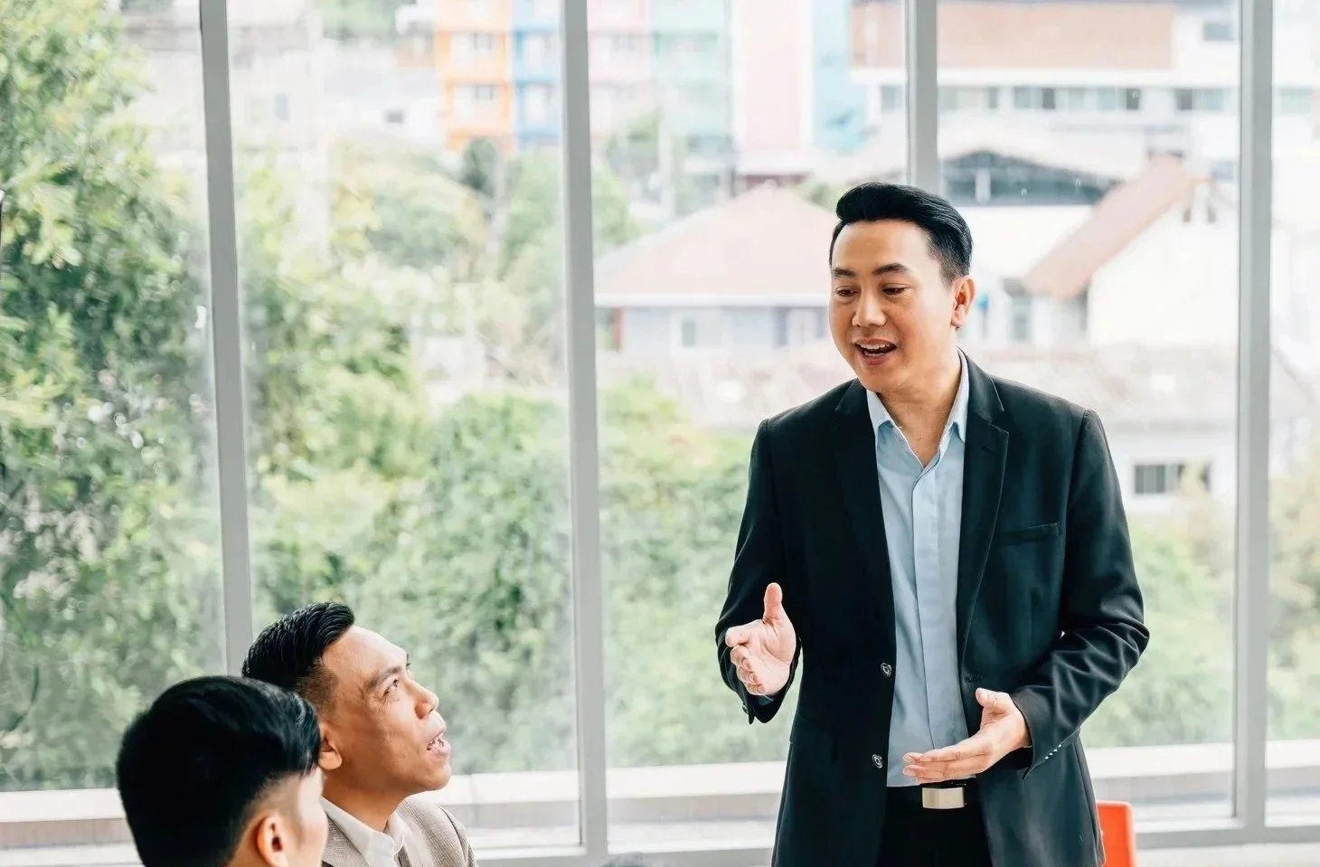 A man in a suit appears to be speaking in a meeting to two other men, seated, in a room with large windows showing trees and buildings outside.