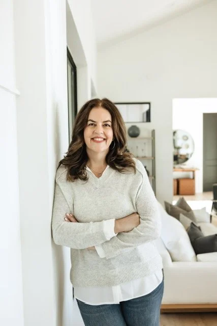 Smiling woman with brown hair wearing a light-colored sweater standing in a modern, well-lit living room.