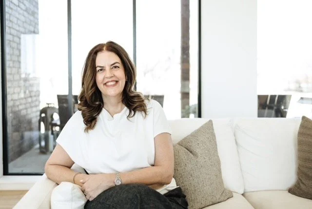 A woman with shoulder-length brown hair, smiling, wearing a white short-sleeve top, sitting on a beige couch with beige pillows in a bright room with large windows.