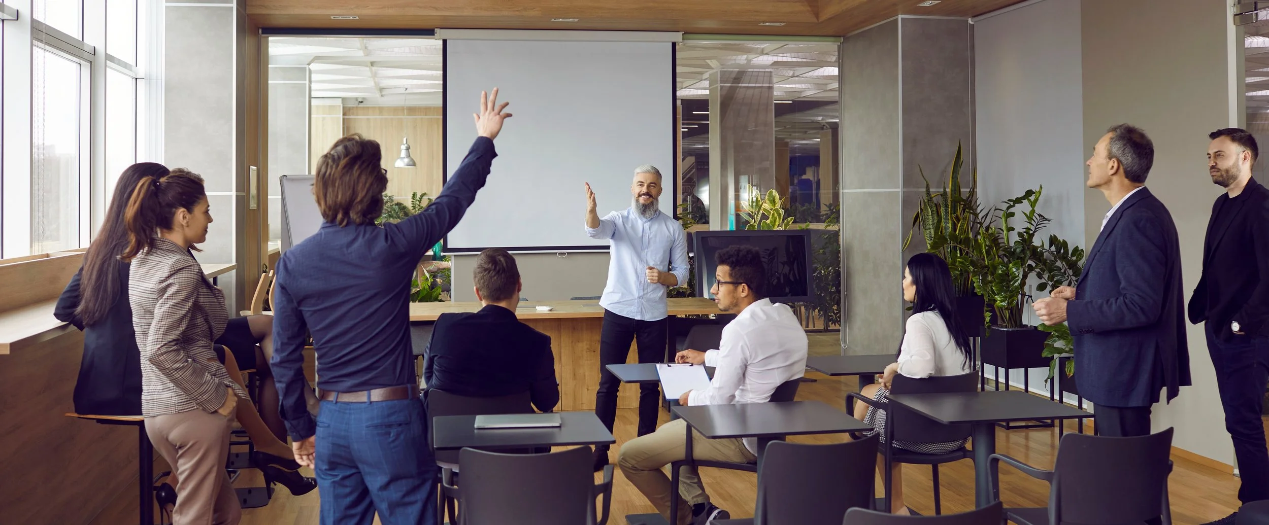 A diverse group of professionals participating in a meeting or presentation in a modern office conference room, with some standing and others seated, a presenter at the front near a large projection screen.
