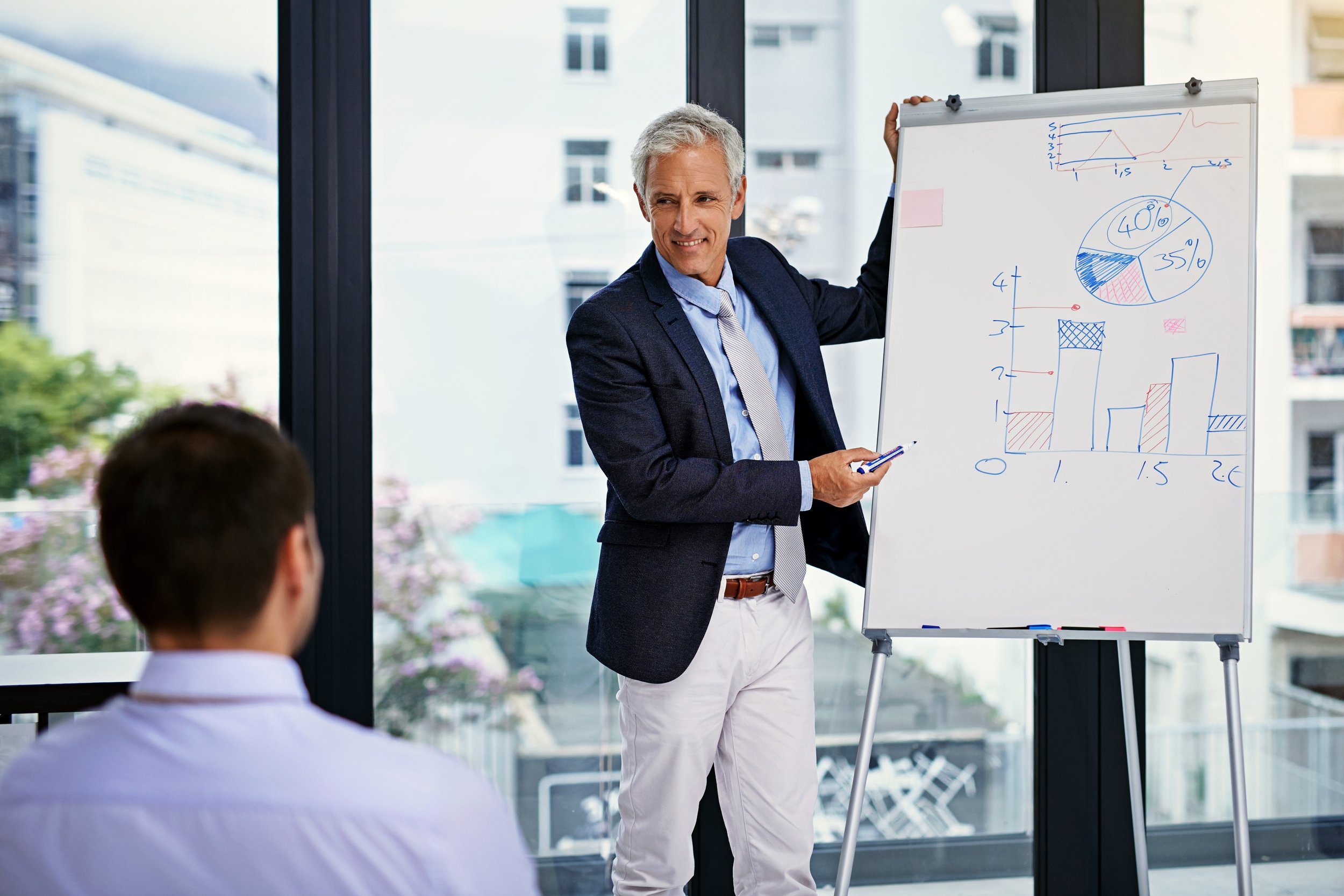 A man in a dark suit and a light blue shirt presenting with a whiteboard filled with charts and graphs in a conference room with large windows.