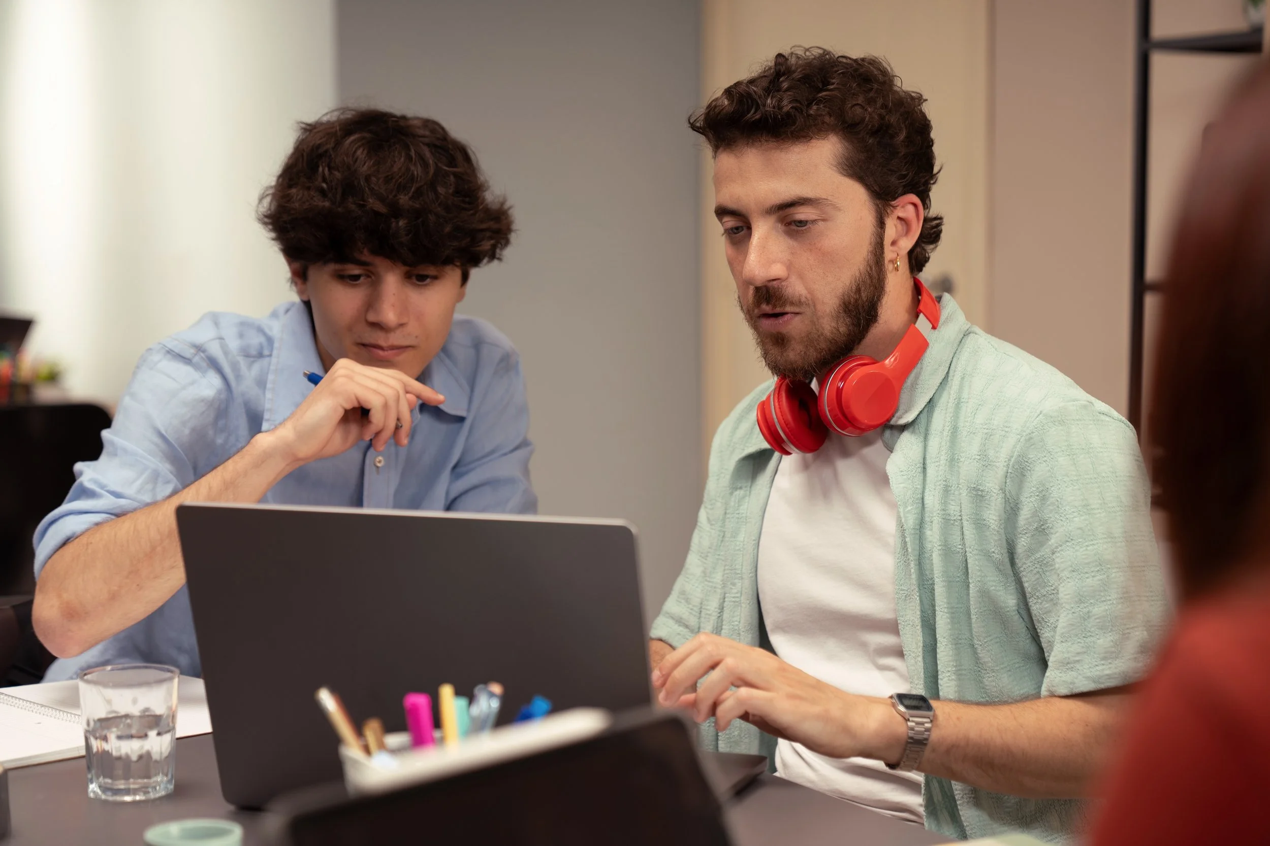 Two men are working together at a desk, focused on a laptop, with a glass of water and office supplies nearby.