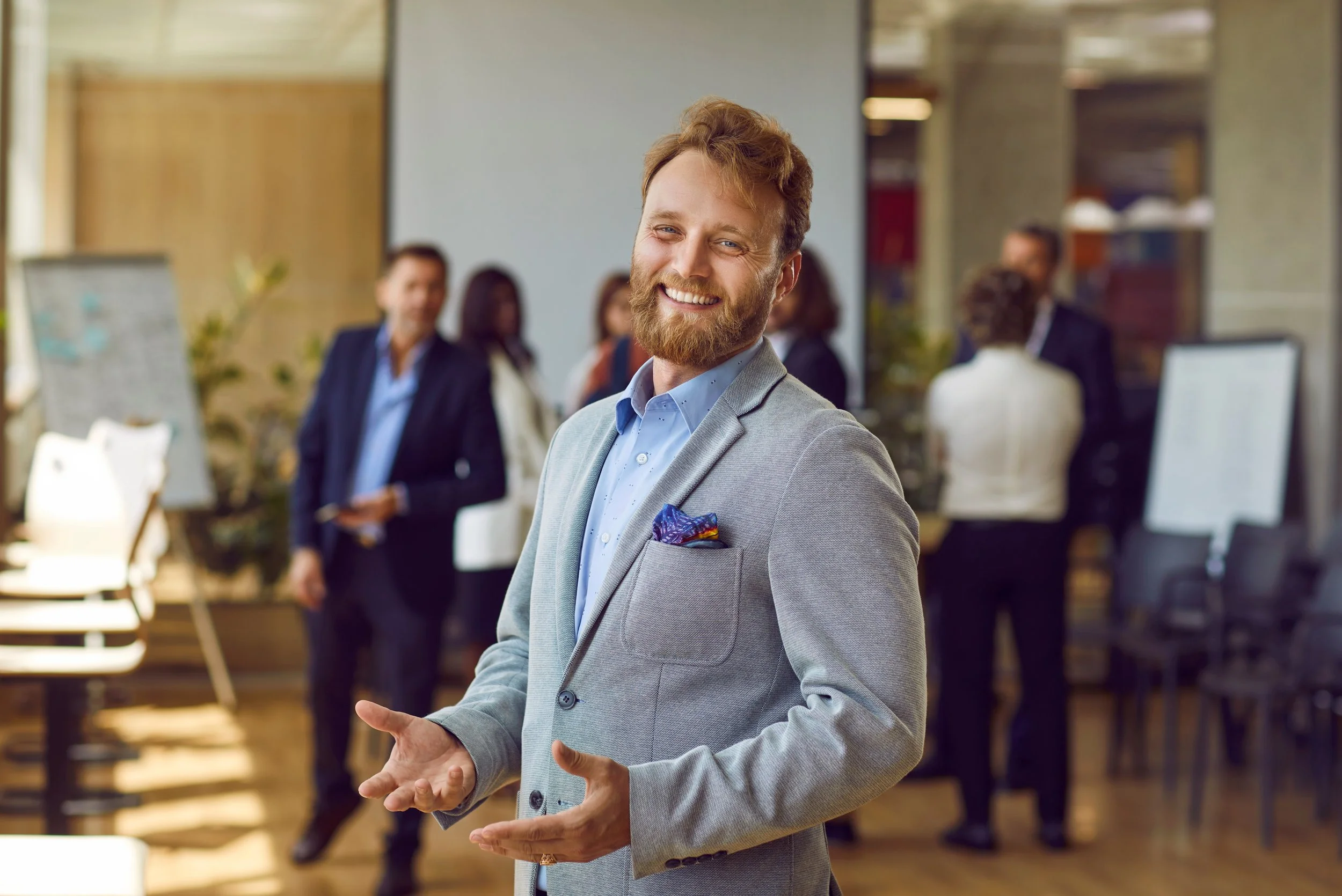 A man with a beard and light brown hair, dressed in a gray blazer and light blue shirt, smiling in a modern office space with several colleagues in the background