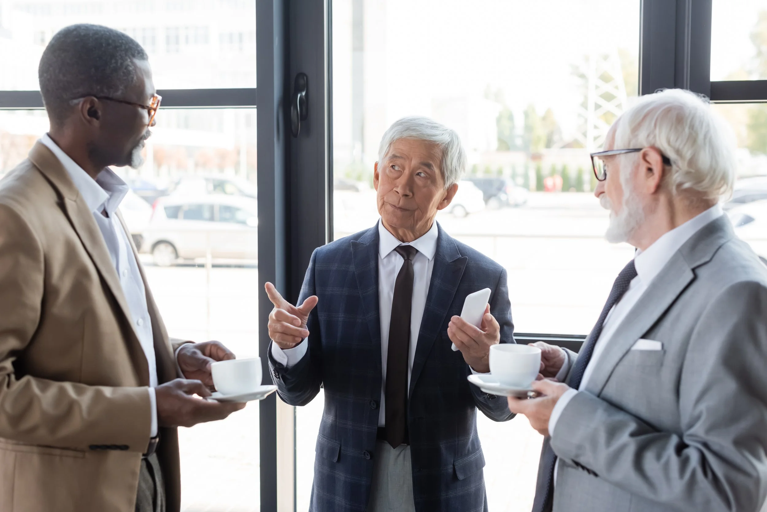 Four men in business attire having a conversation over coffee in a modern office with large windows.