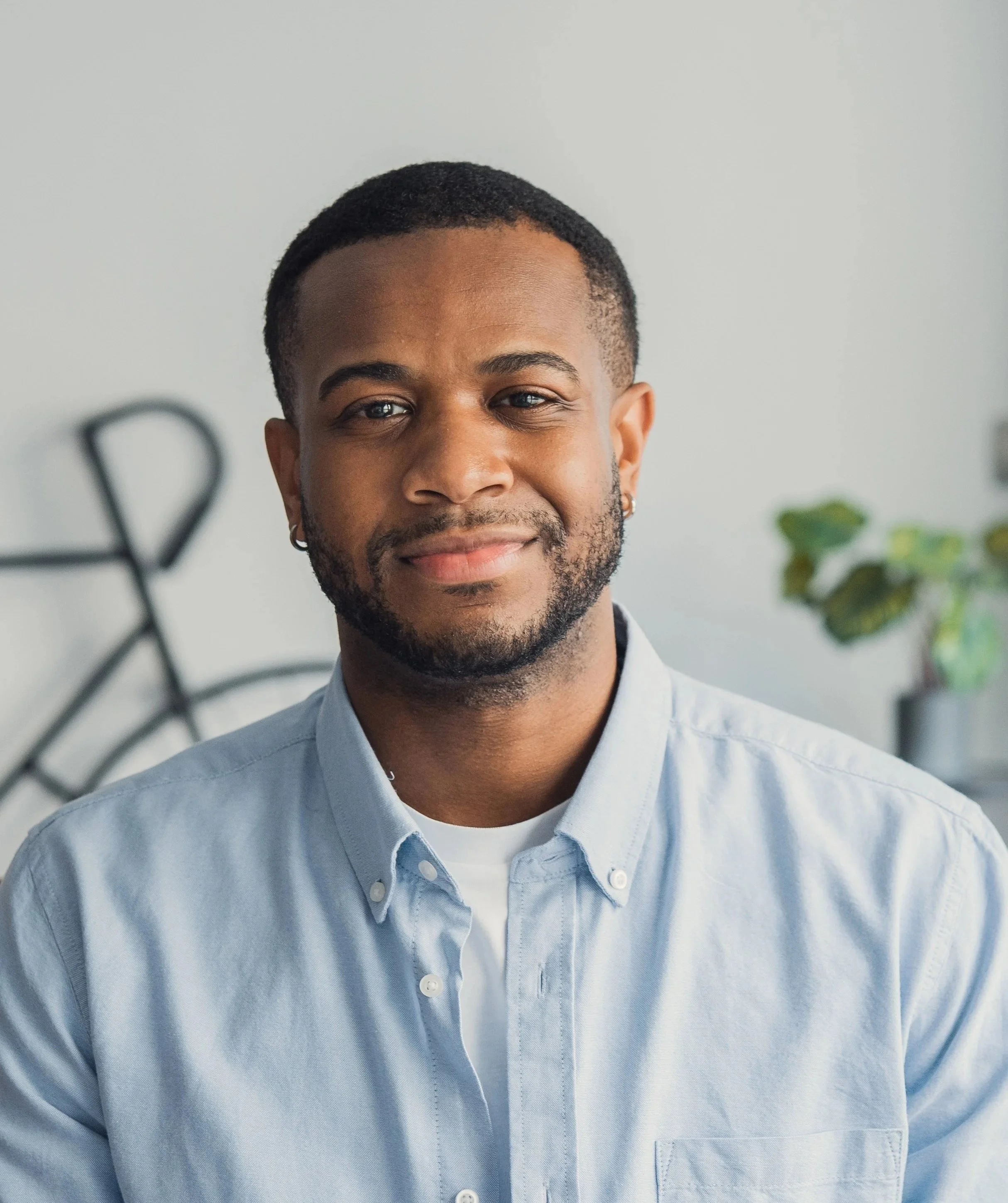 Close-up of a smiling young African American man with short curly hair, wearing a light blue button-up shirt, in a well-lit indoor setting with blurred plants in the background.