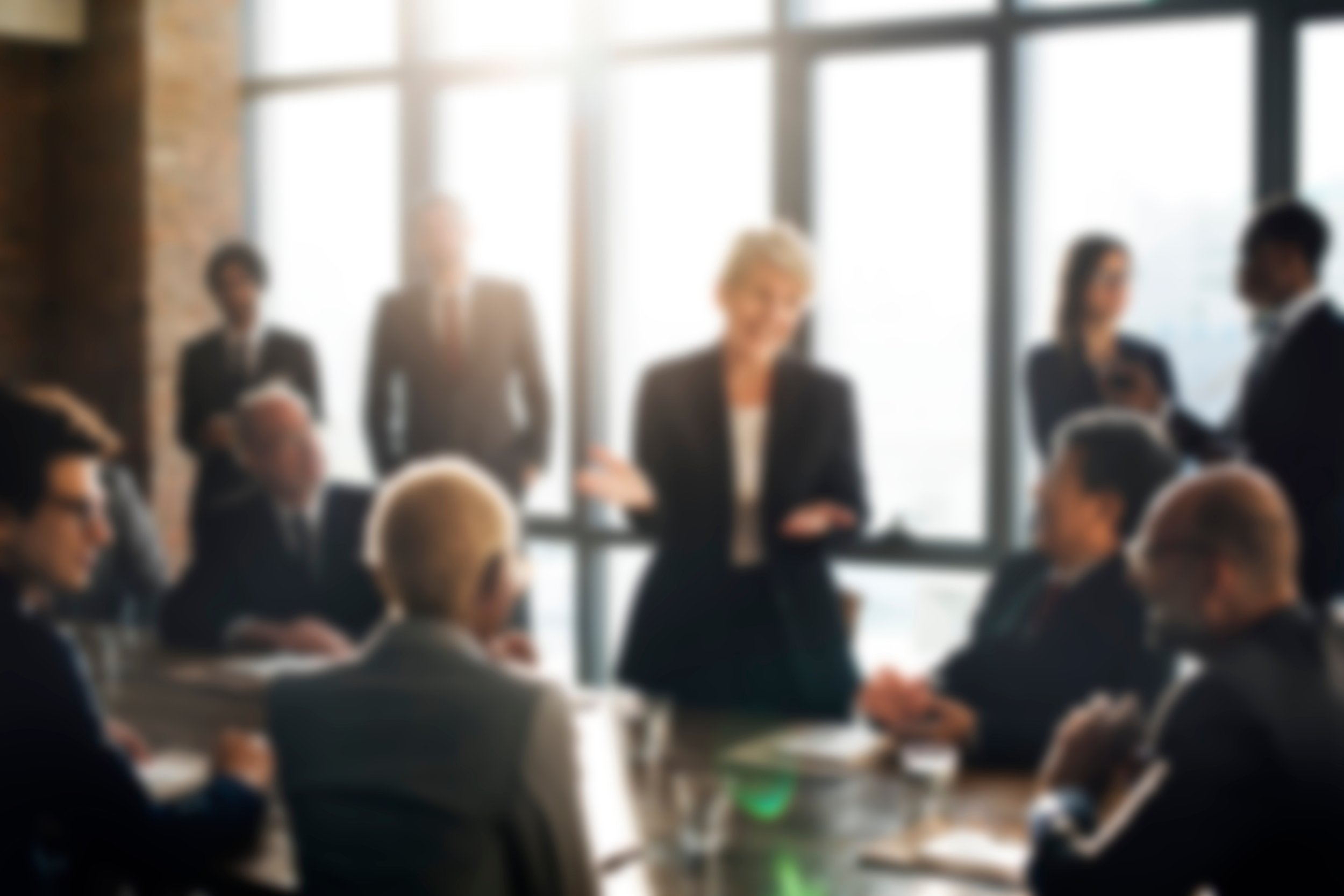 Blurred image of people in a business meeting or conference room, with several individuals sitting at tables and a few standing, in front of large windows.