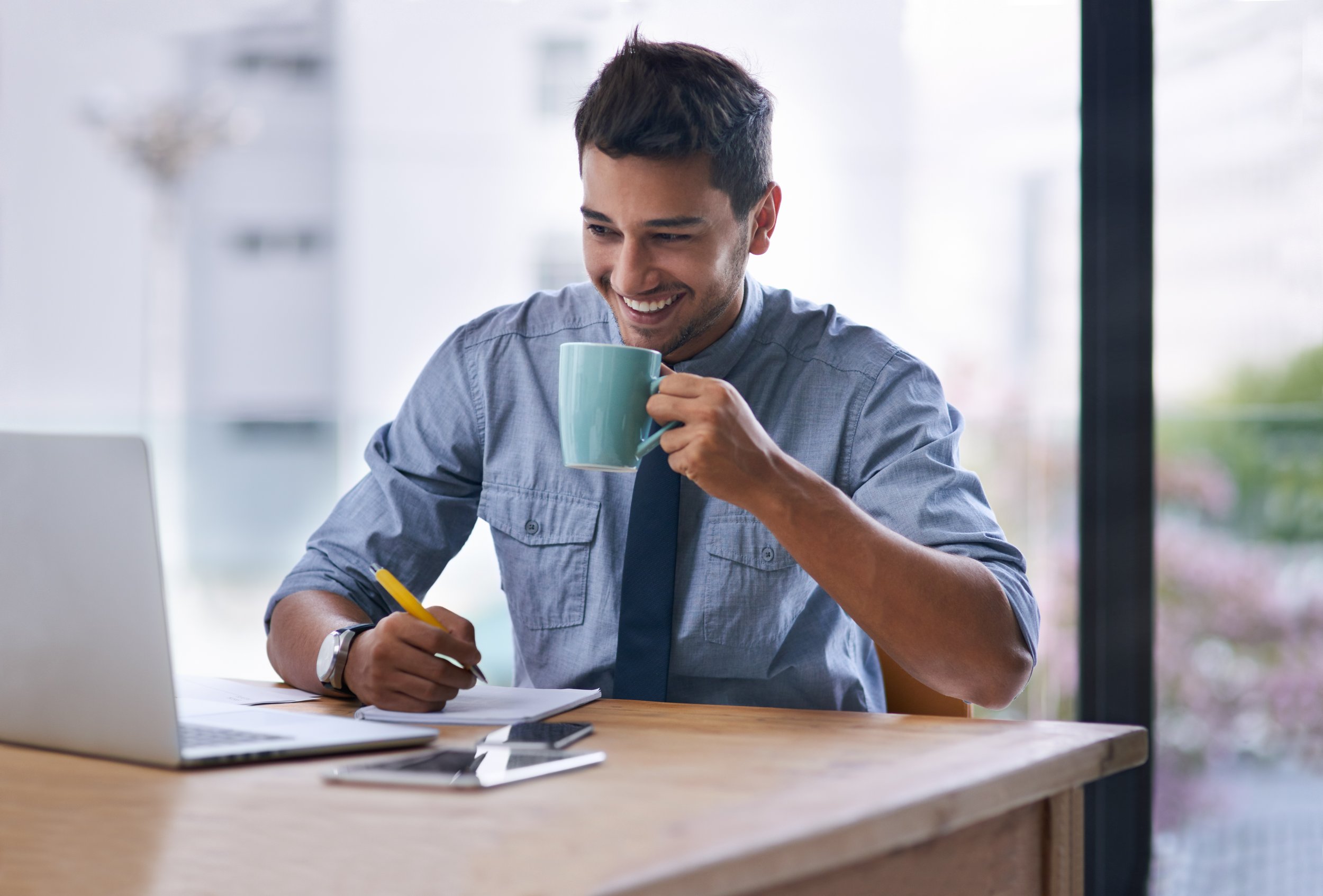 A young man with short dark hair and a beard, wearing a blue button-up shirt, sitting at a wooden desk in front of a laptop, smiling while holding a light blue mug and writing with a yellow pen. There are papers, a smartphone, and a tablet on the desk. Large windows in the background reveal a blurred cityscape.